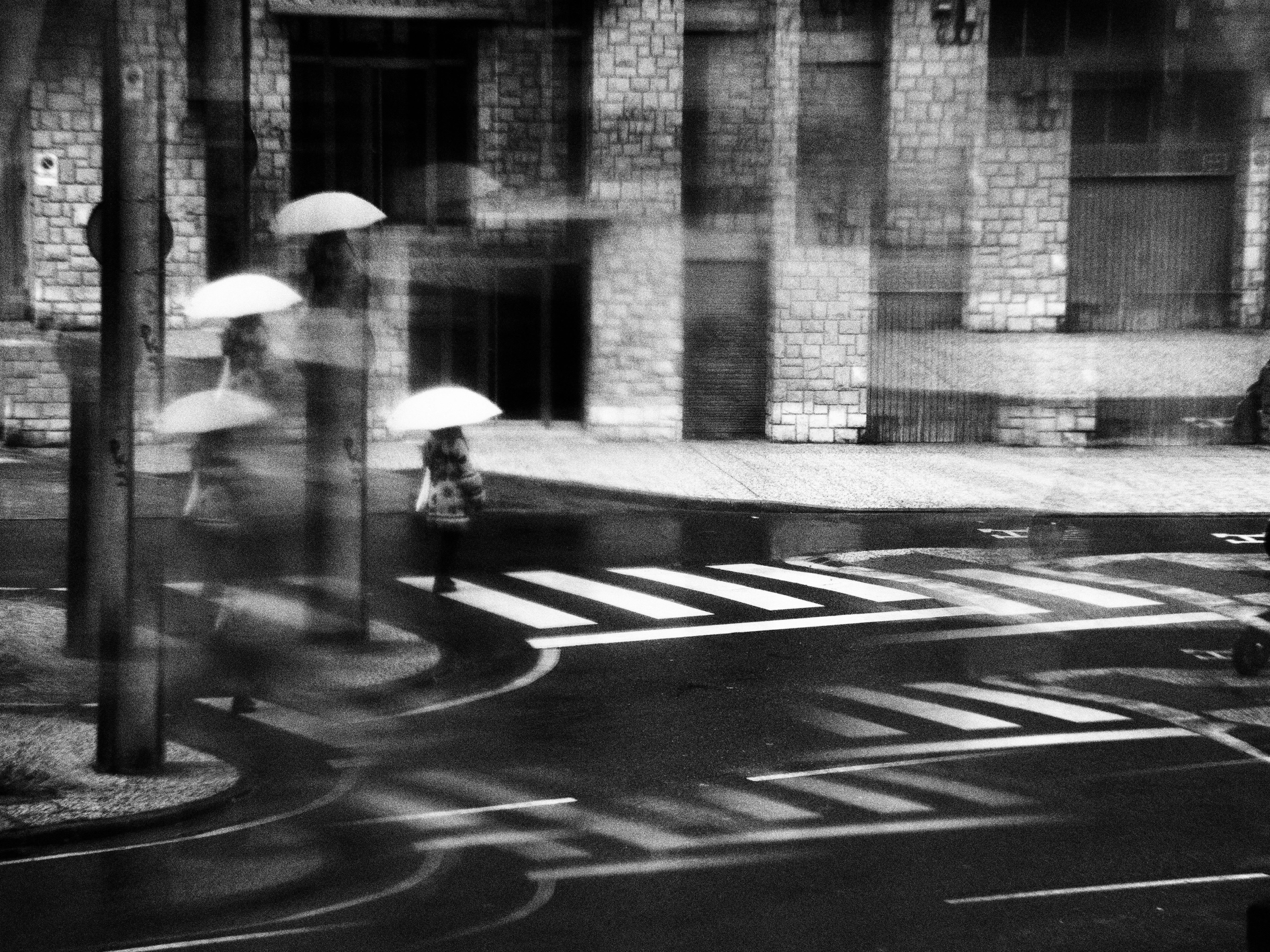 Black and white city street with motion blur and umbrellas crossing a rainy intersection.