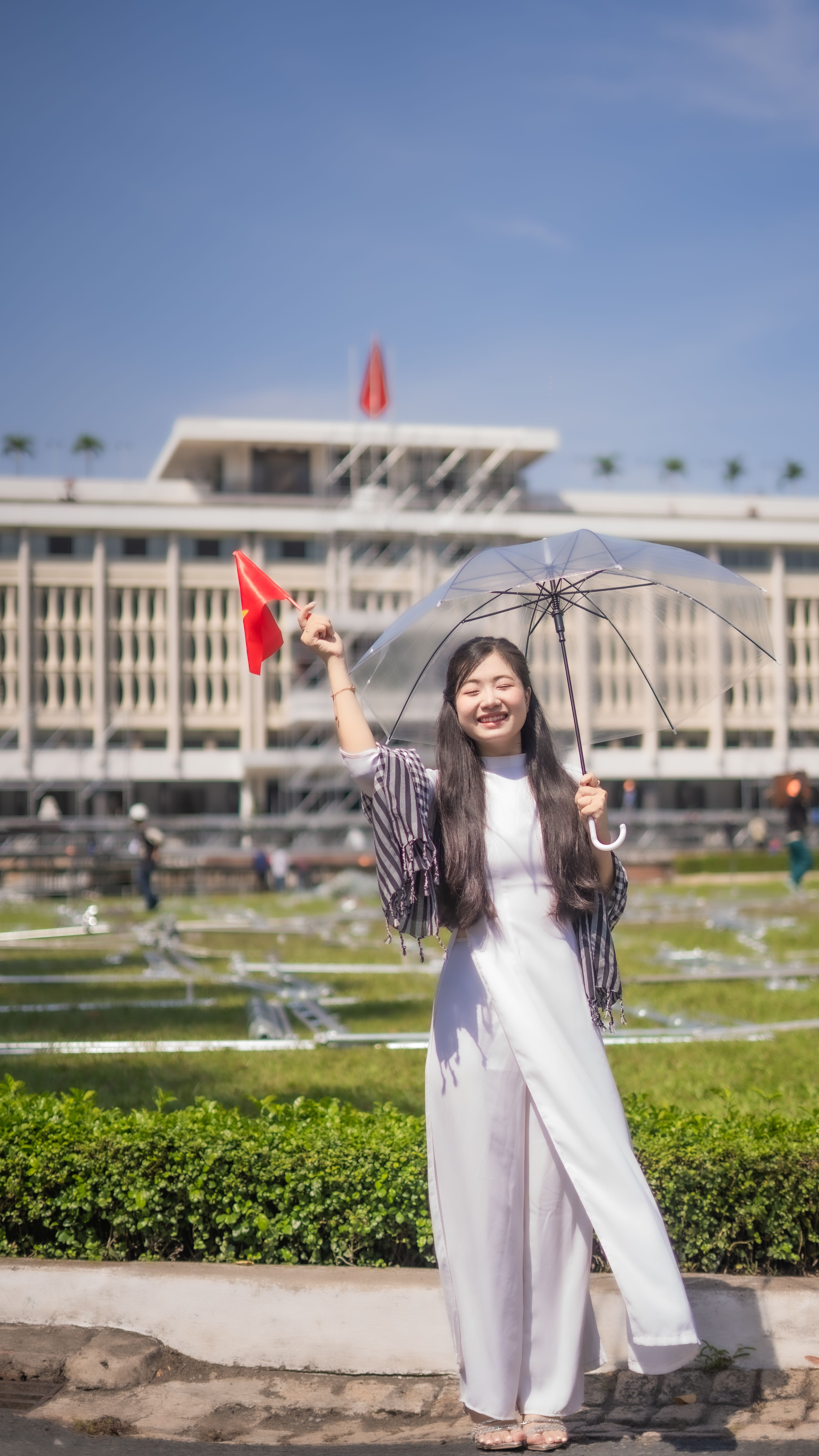 Smiling woman with umbrella and flag in front of a modern landmark on a sunny day.
