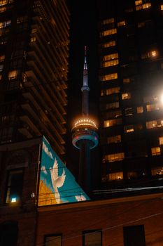 Stunning night view of Toronto's CN Tower framed by illuminated buildings, with a colorful mural in the foreground.