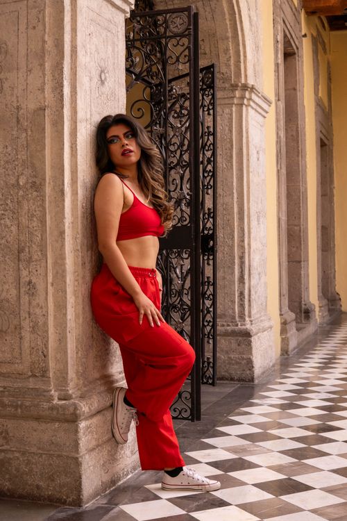 Free Confident woman in a red outfit leaning against a stone wall indoors with checkerboard flooring. Stock Photo