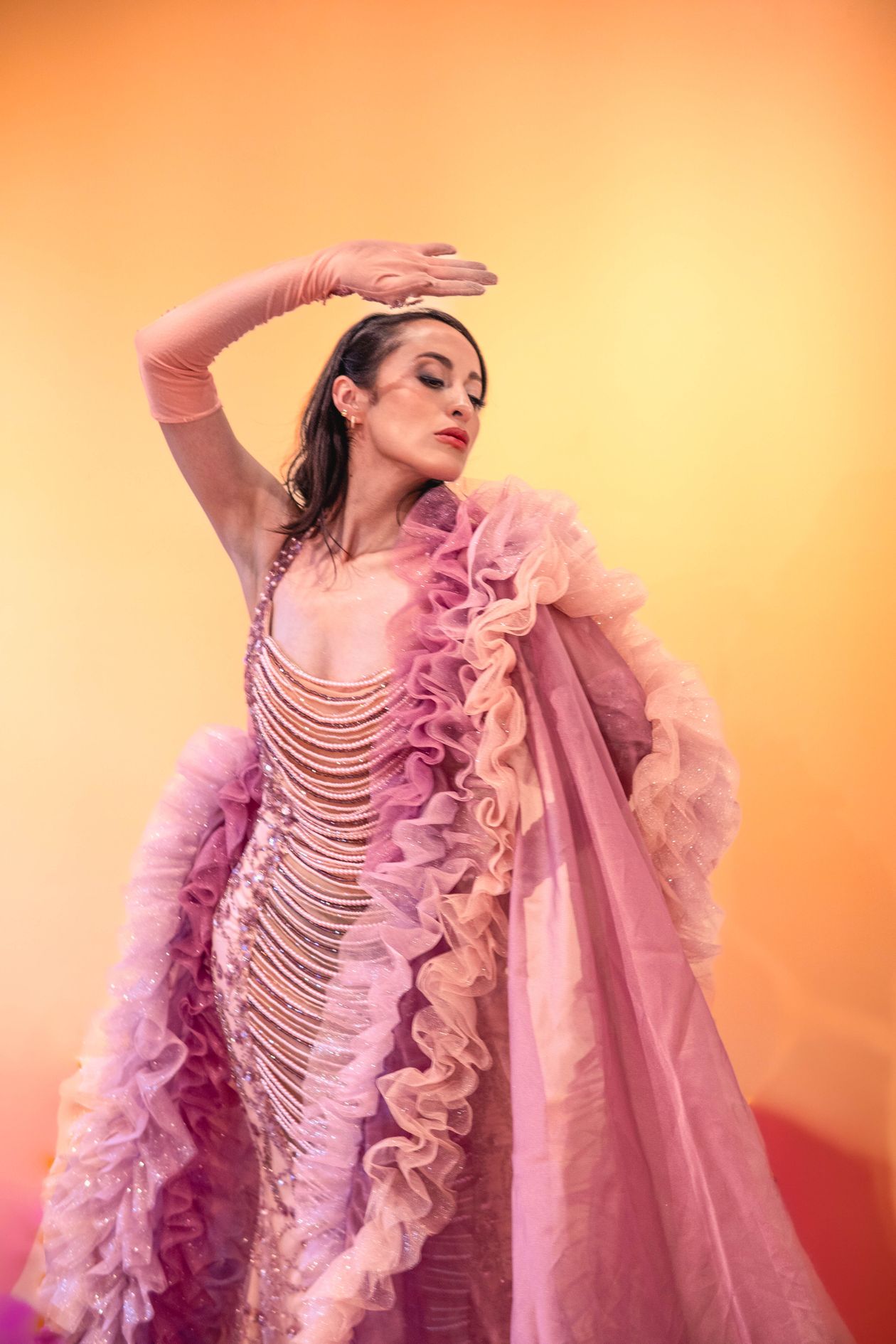 Woman in a pink beaded gown with a ruffled shawl posing against a warm orange backdrop