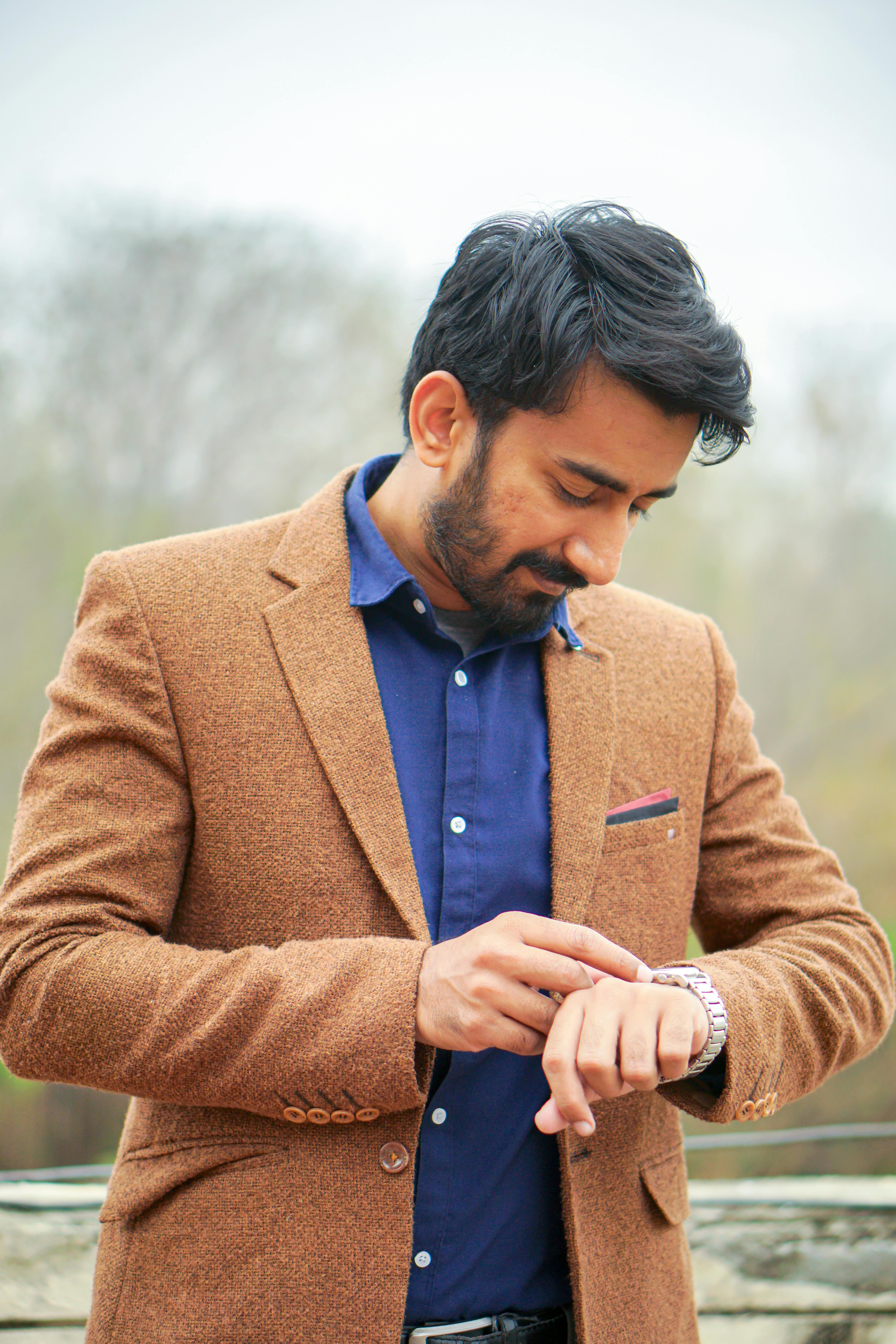 Portrait of a young man in a brown suit looking at his watch, set against an outdoor background in Islamabad, Pakistan.