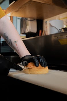 A chef wearing black gloves prepares a burger bun on a cutting board in a restaurant kitchen.
