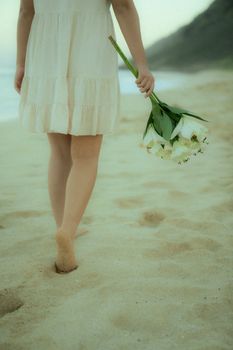 A woman in a white dress walks barefoot on a sandy beach, holding a bouquet of white flowers.