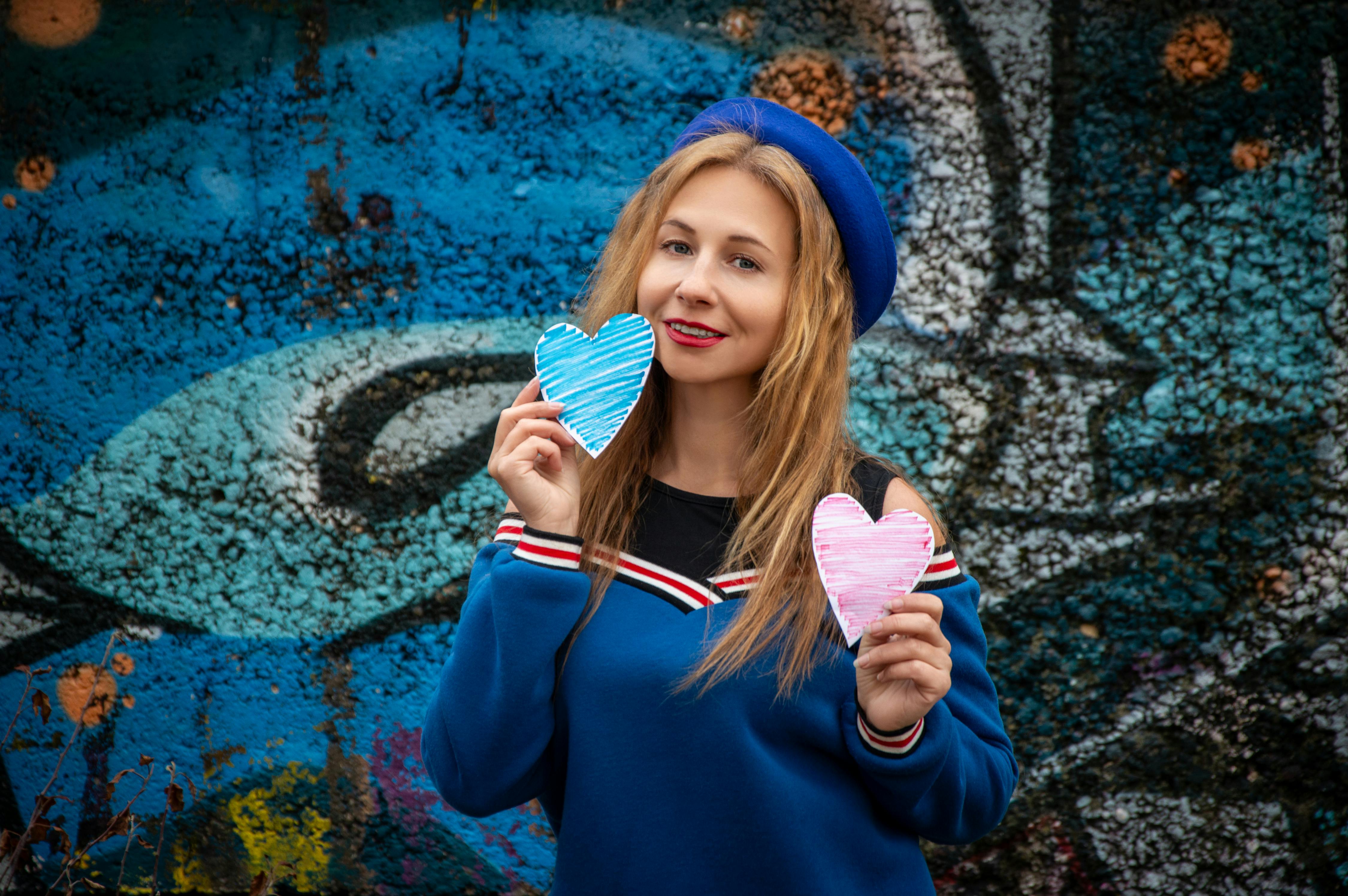 Smiling woman in blue outfit holding paper hearts against colorful graffiti background.