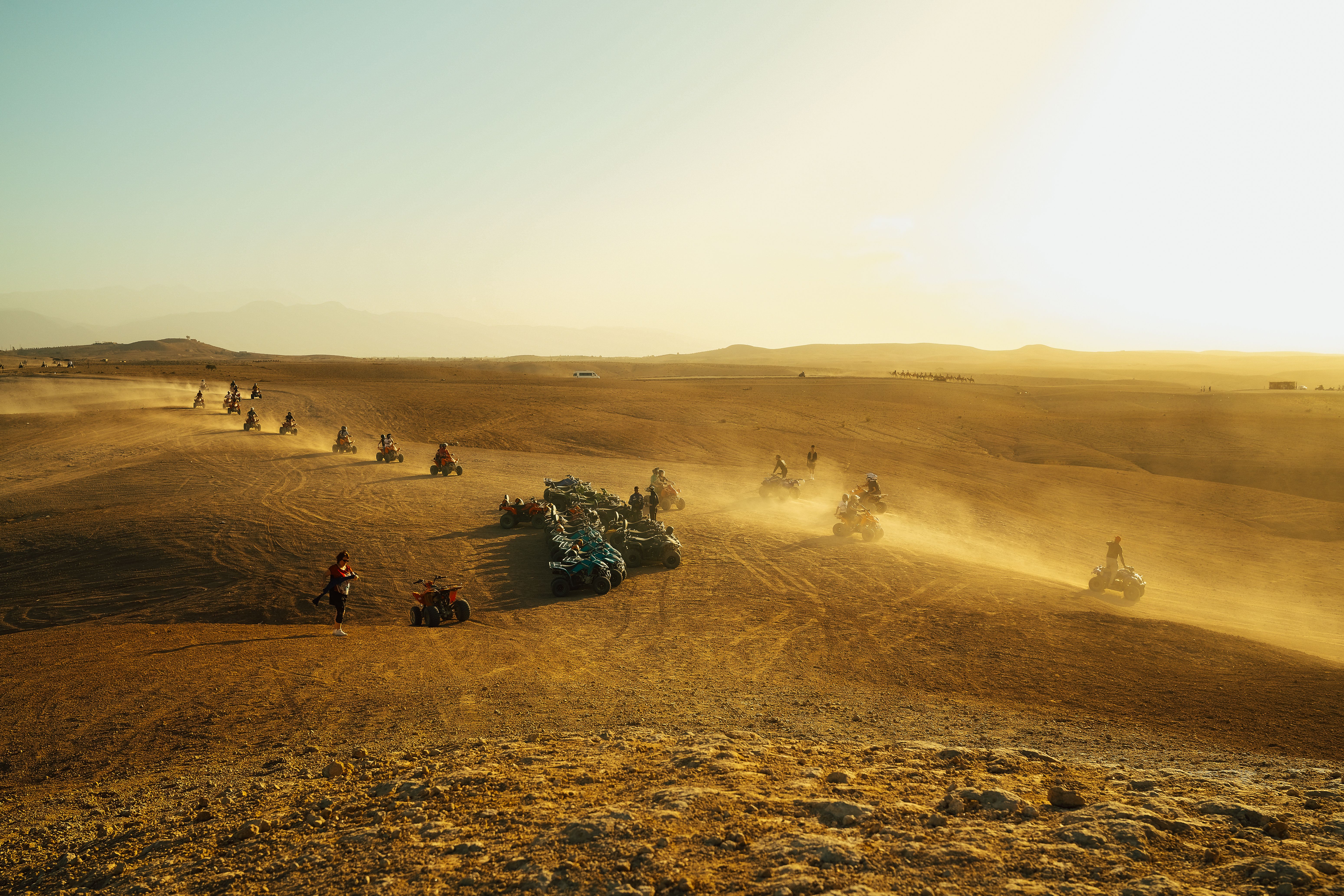 Group of people riding quad bikes across a vast, sandy desert terrain at sunset.