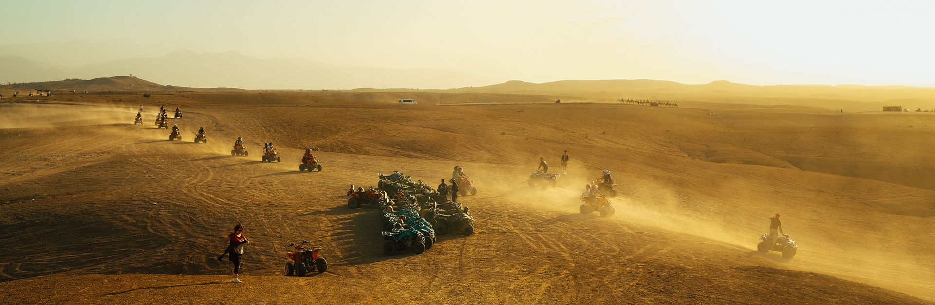 Group of people riding quad bikes across a vast, sandy desert terrain at sunset.