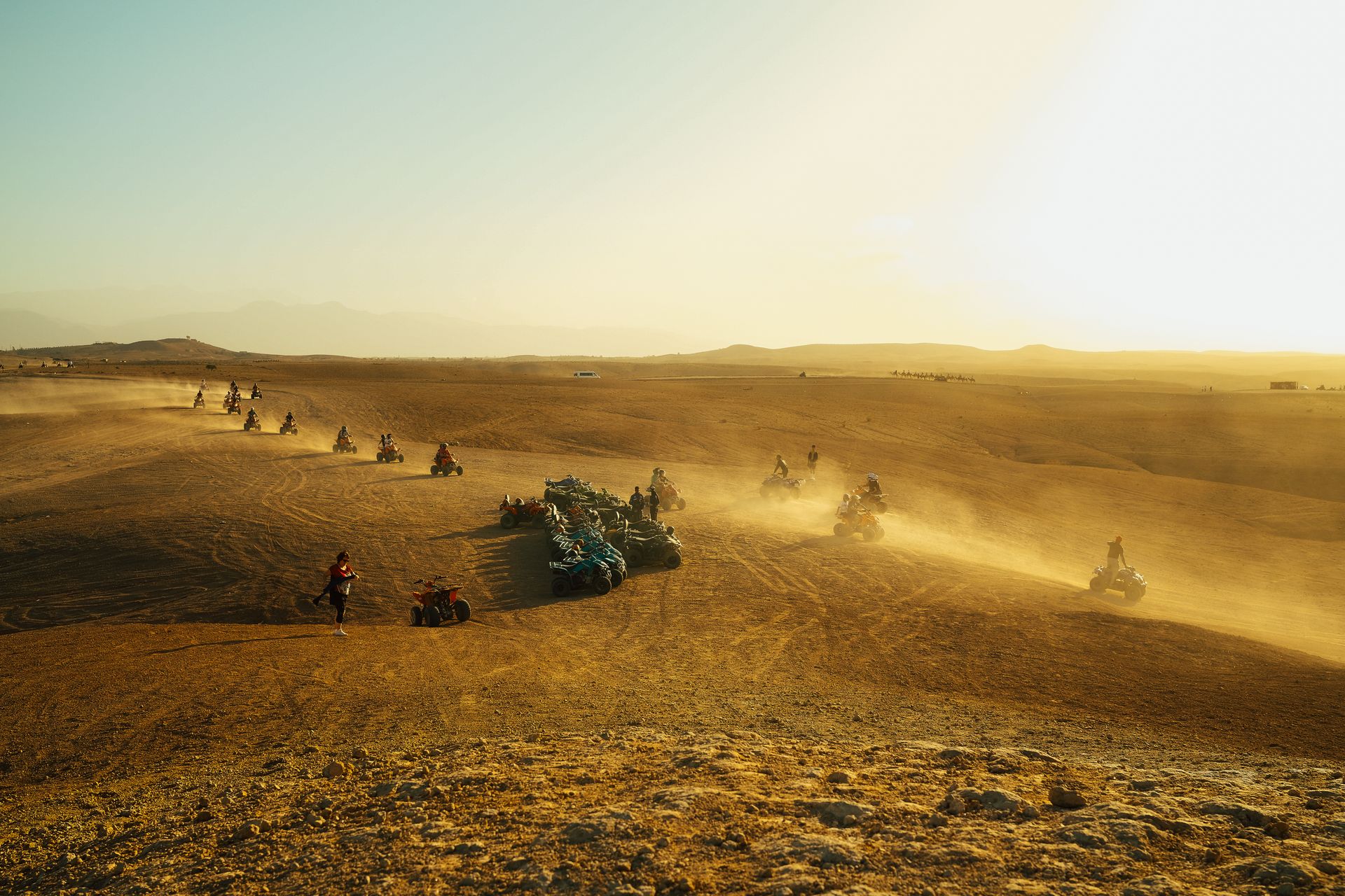 Group of people riding quad bikes across a vast, sandy desert terrain at sunset.