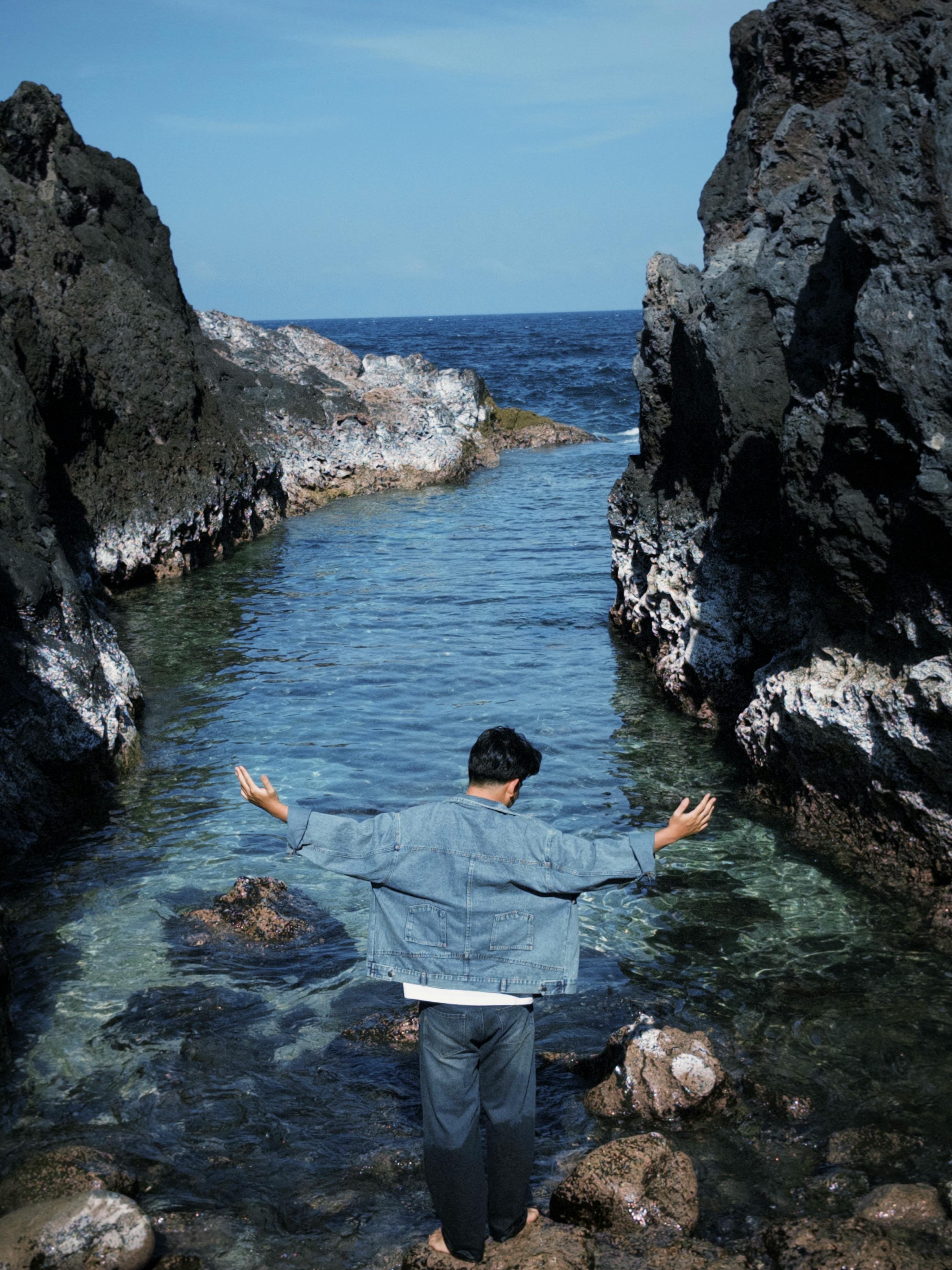 Man stands amidst rocky cliffs enjoying the serene coastal view, arms outstretched.