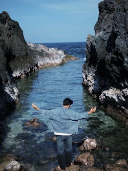 Man stands amidst rocky cliffs enjoying the serene coastal view, arms outstretched.