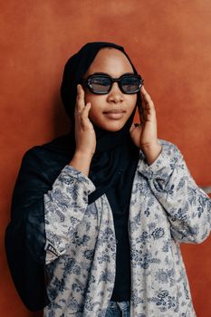 Confident woman in floral outfit, wearing sunglasses and hijab against a textured wall.