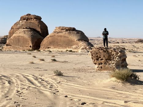 A lone person stands on sandstone with desert formations in the background under a bright sky.