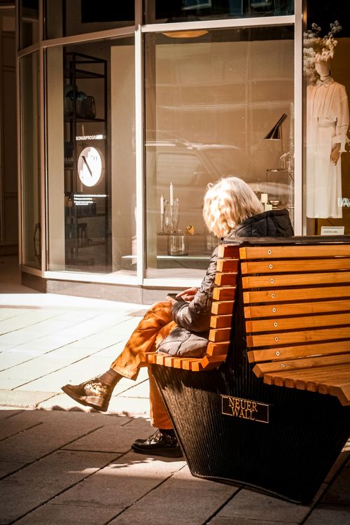 Free Elderly woman enjoying sunlight while resting on a bench in a city shopping area. Stock Photo