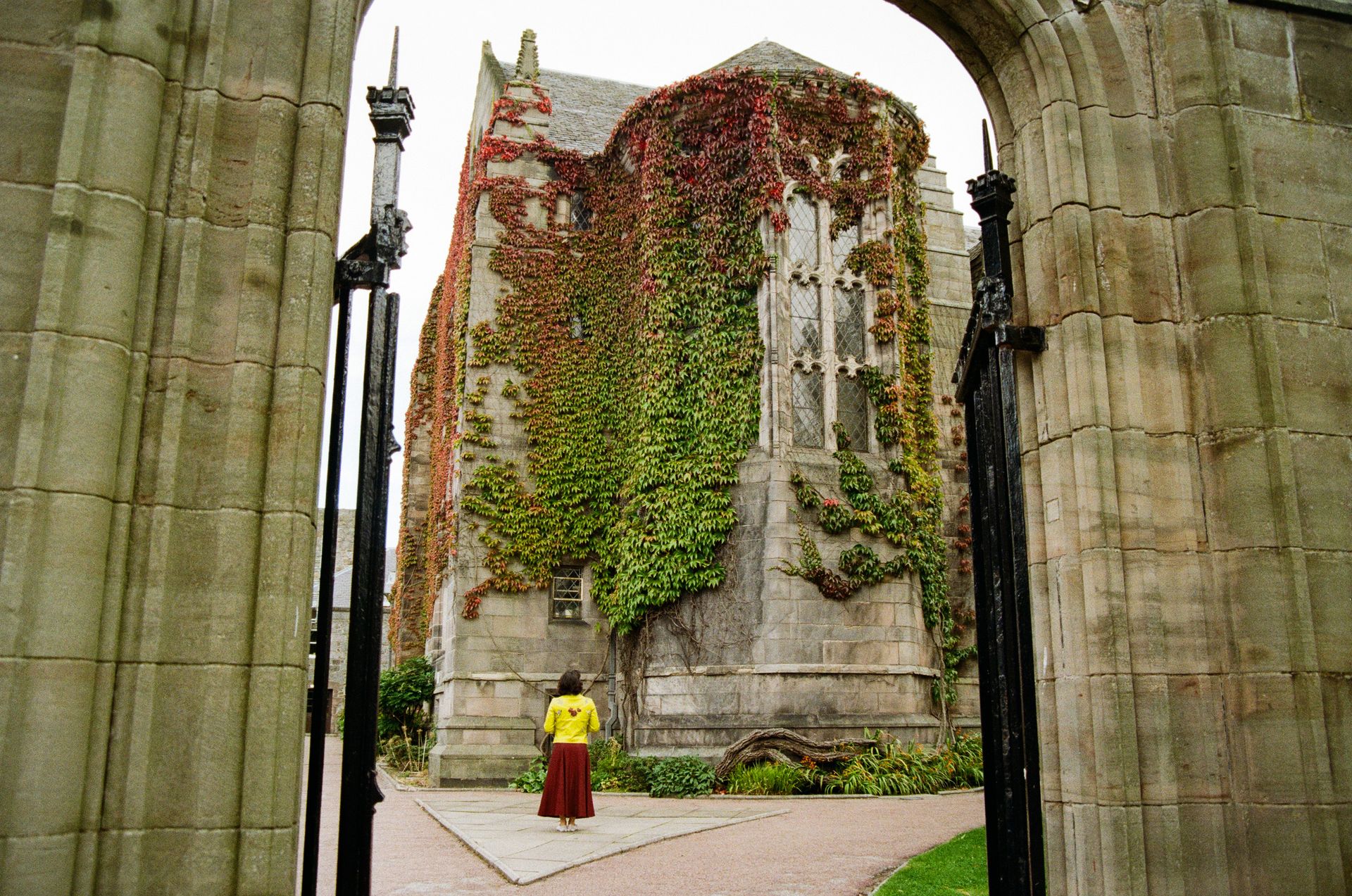 A lone woman stands by an ivy-covered historic building in Aberdeen, captured through an archway.