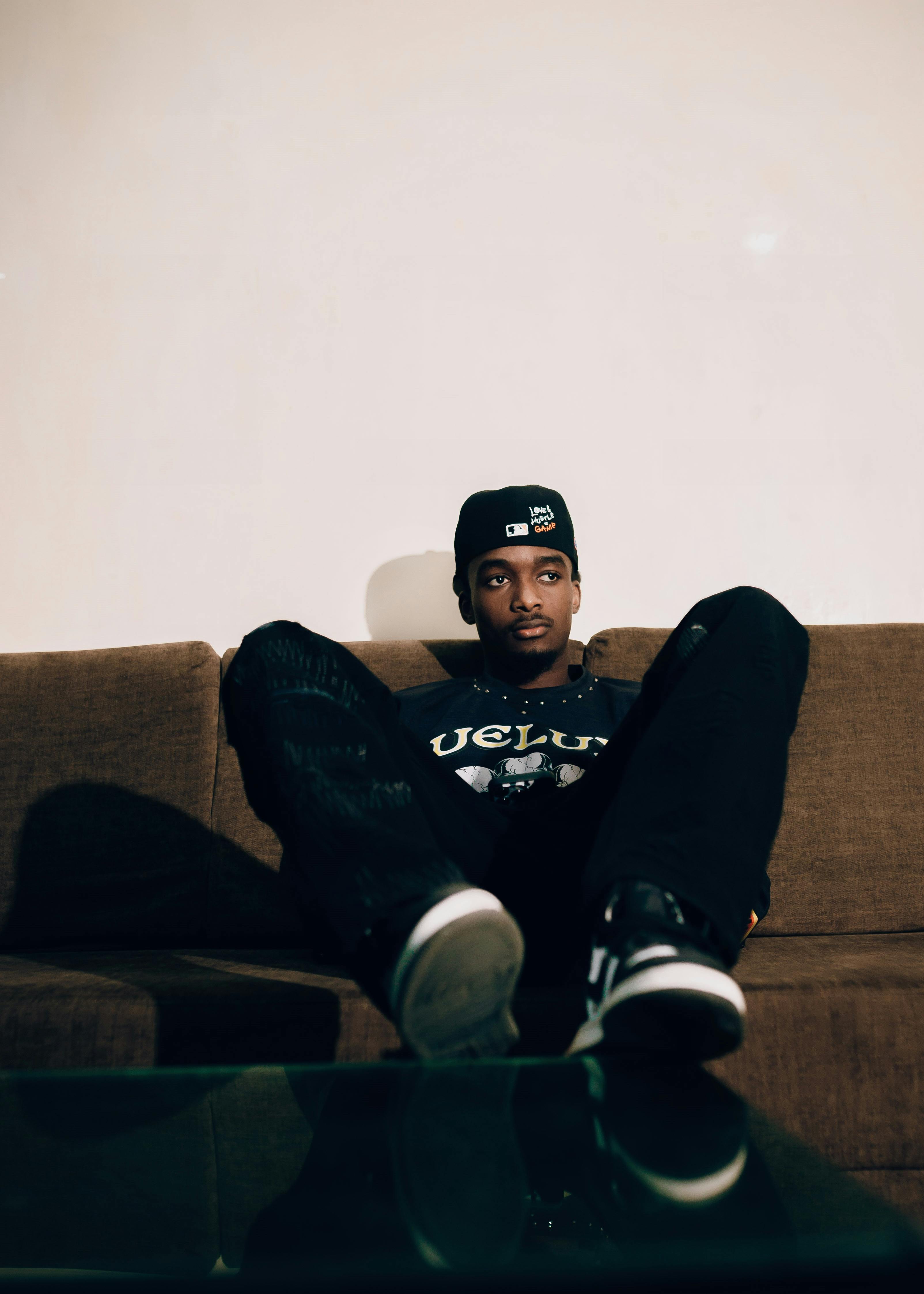 Young man relaxing on a sofa indoors, wearing casual attire with a cap.