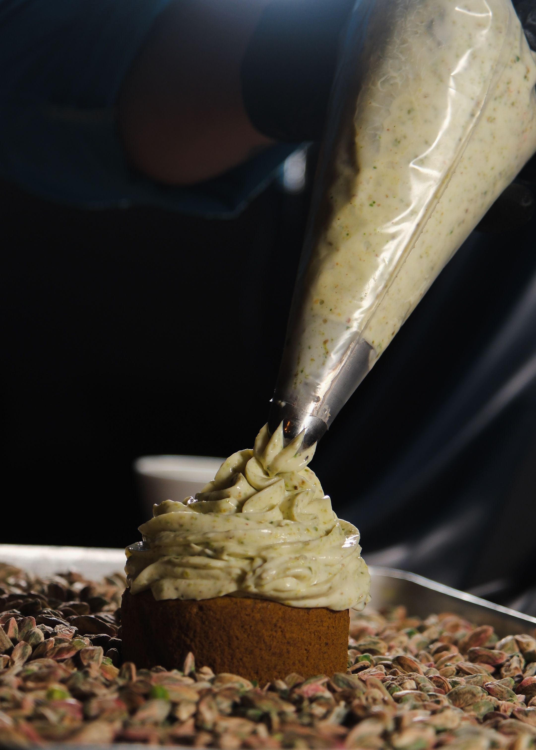 Hand decorates a pistachio cake using frosting, surrounded by whole pistachios.