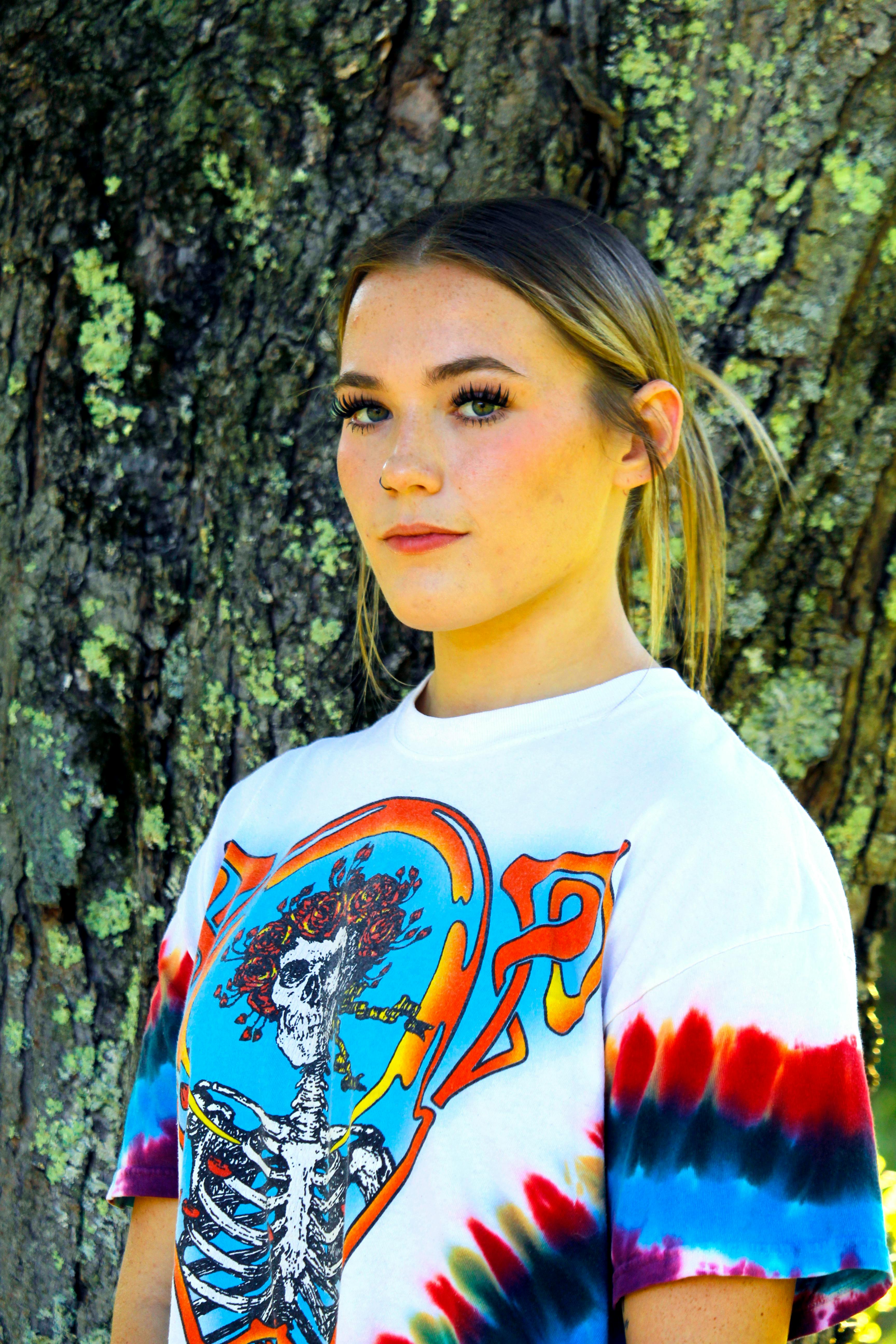 Portrait of a young woman in a colorful tie-dye shirt posing against a tree outdoors.