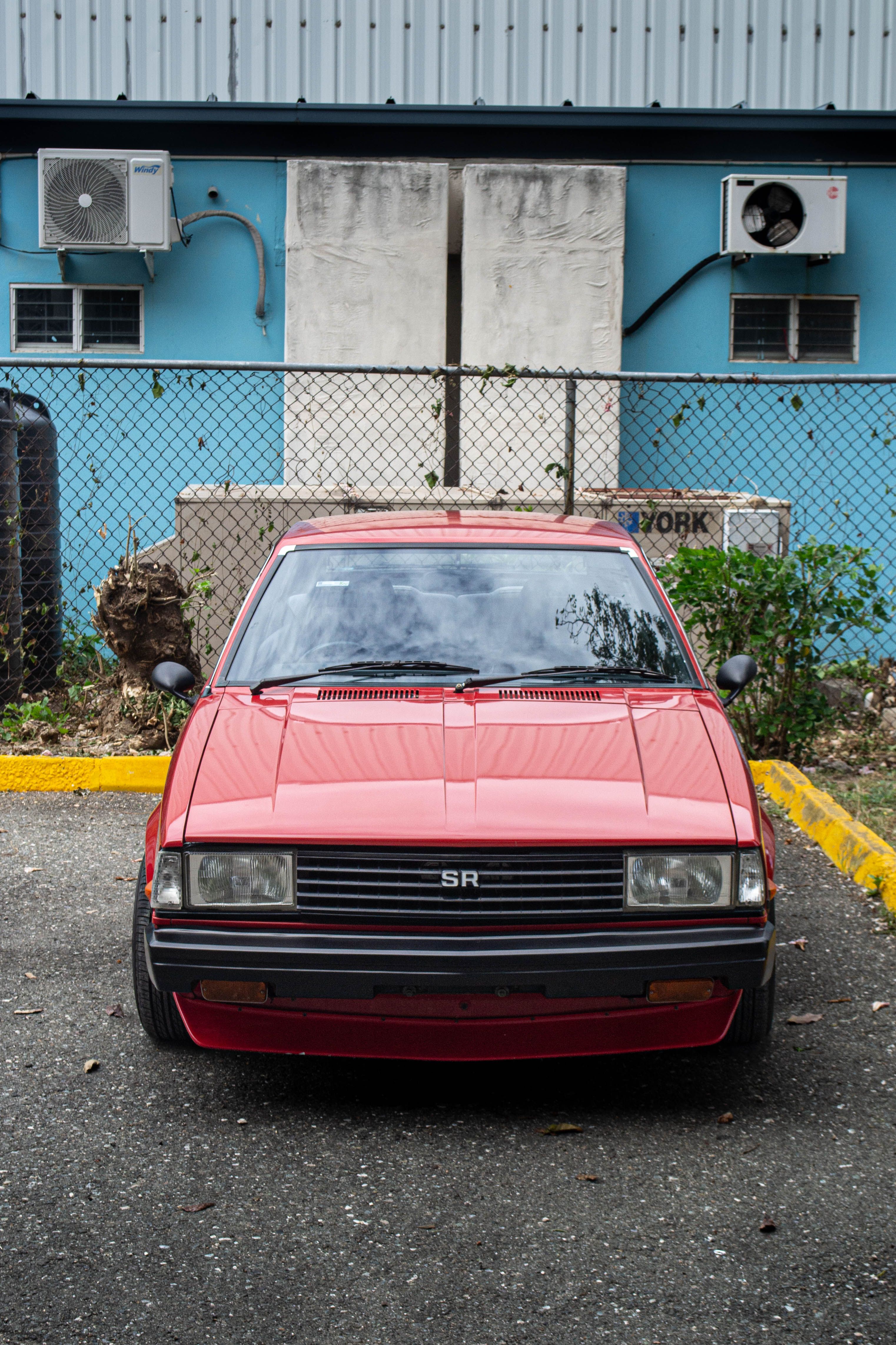 Front view of a vintage red car parked outdoors in an urban area, featuring a chain-link fence and utility boxes.