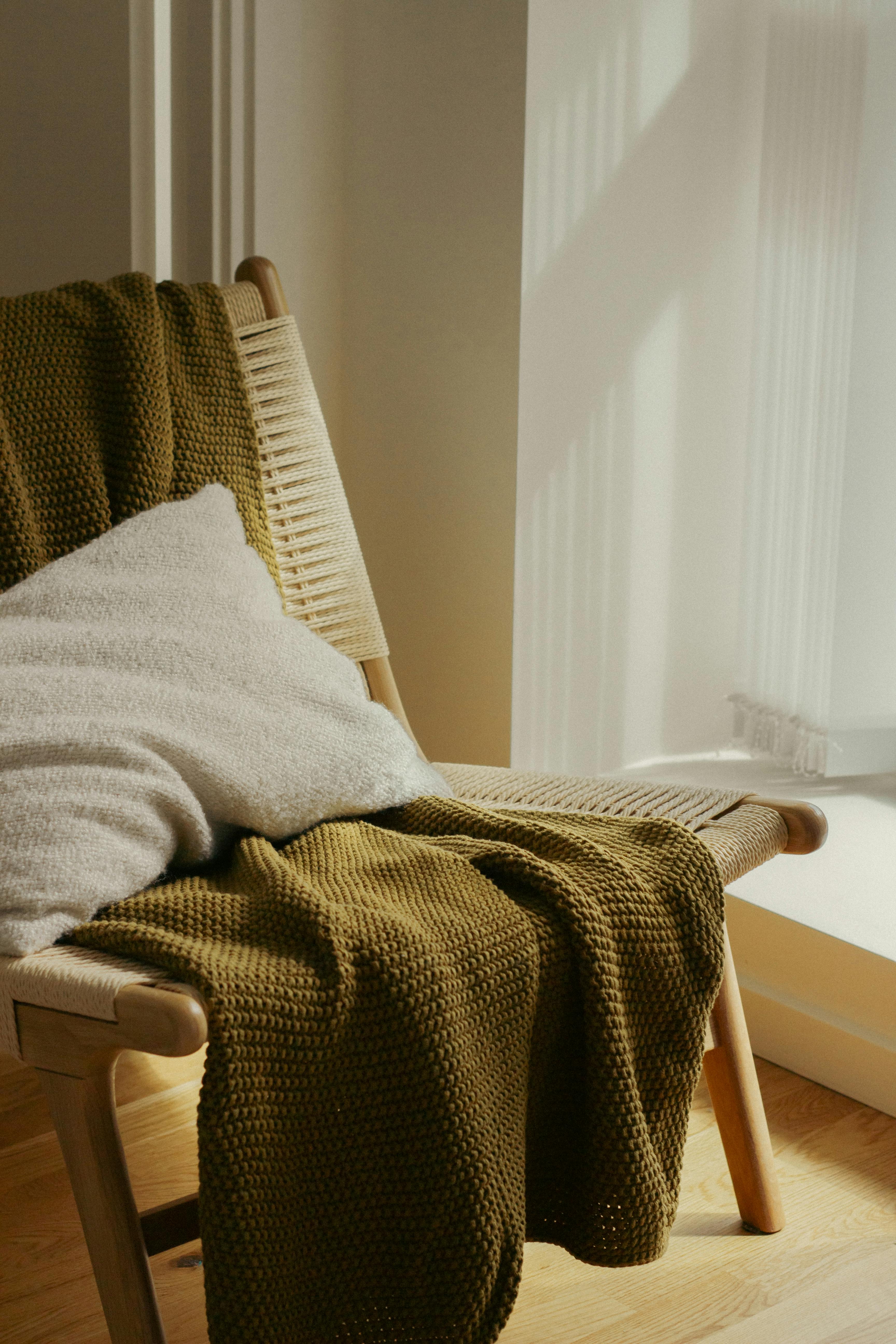 Free A stylish chair with green blanket and pillow in sunlit room, perfect for cozy interiors. Stock Photo