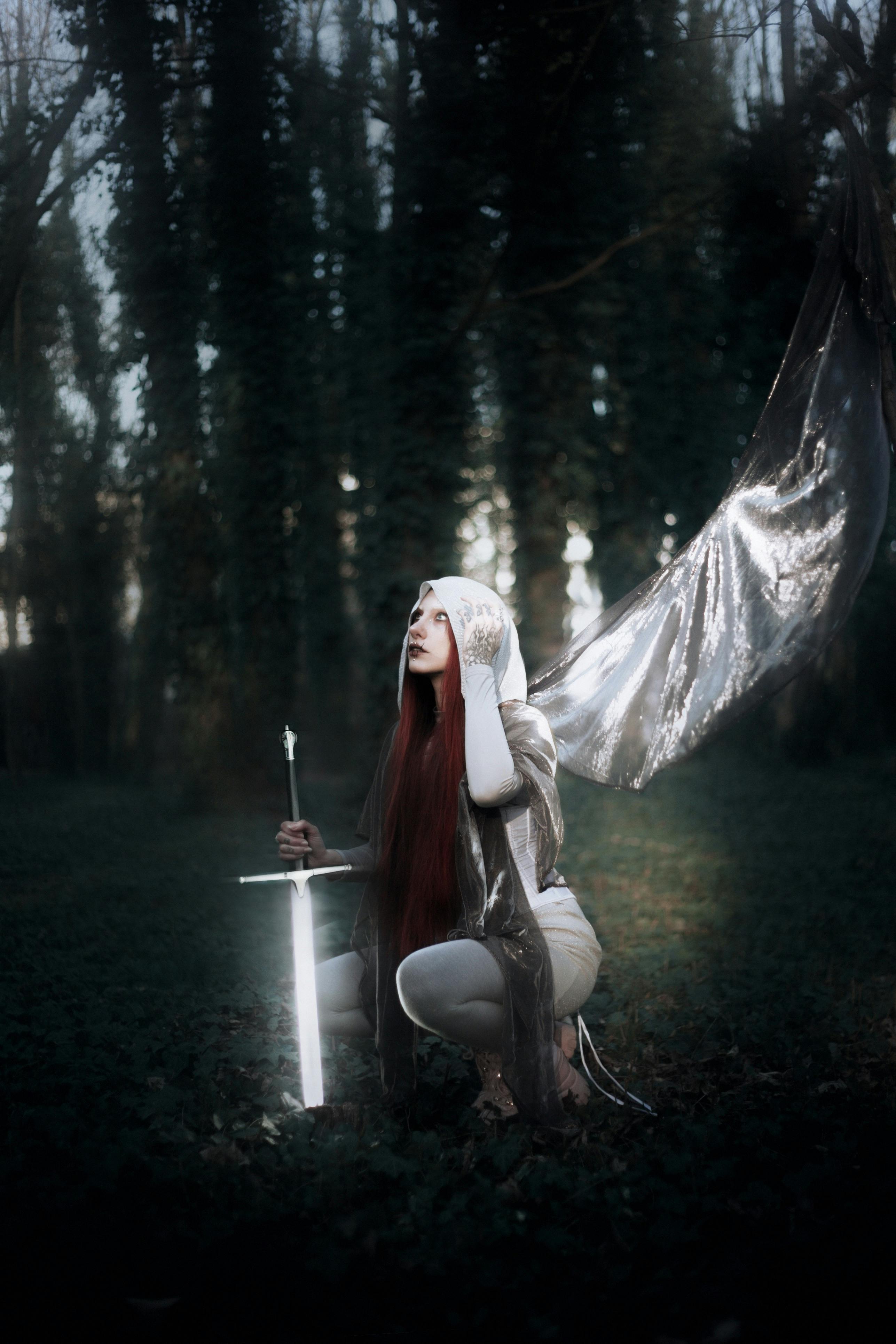 An ethereal angelic figure kneels amidst a shadowy forest, holding a glowing sword.