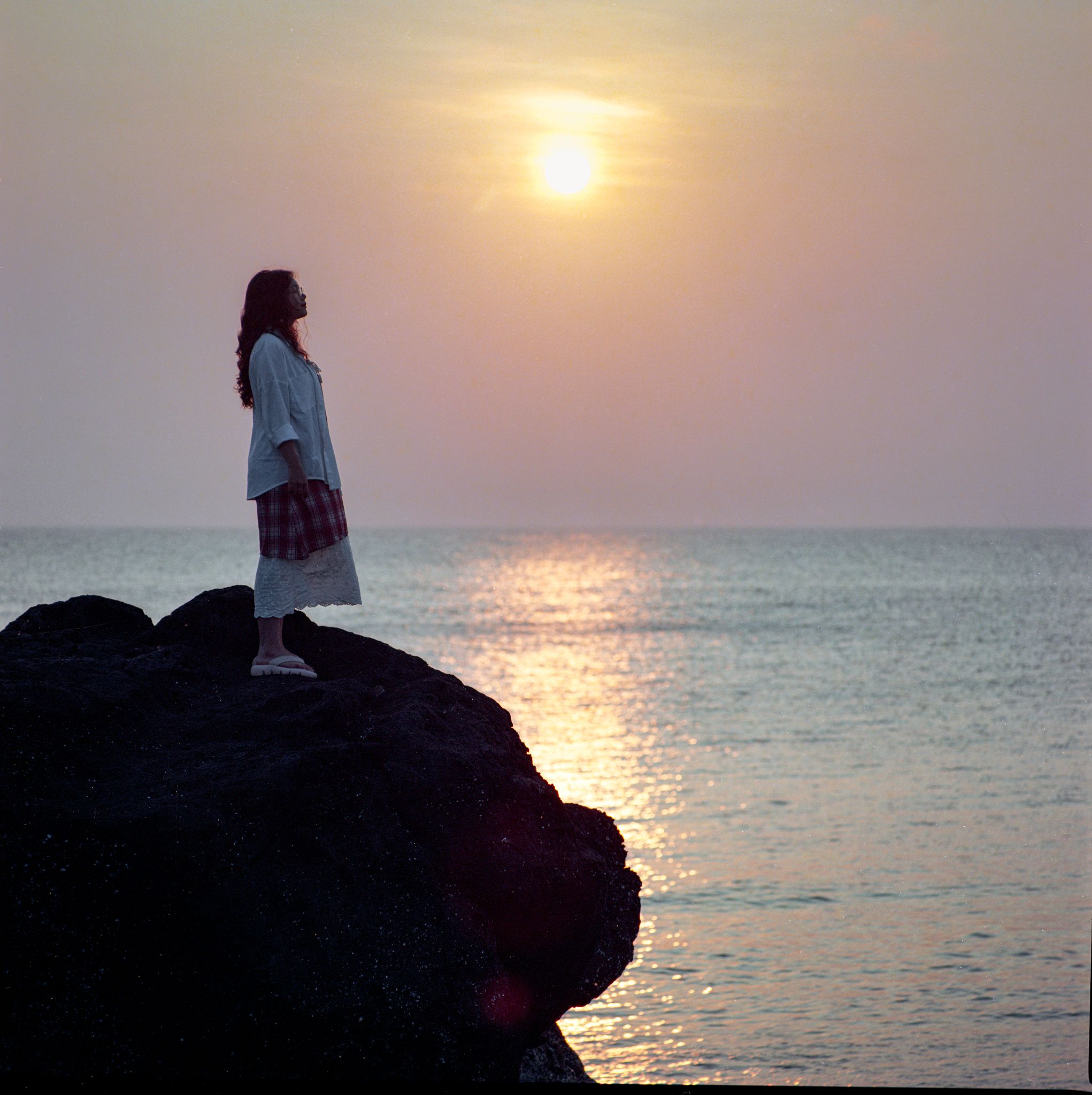A woman stands on a rocky shore in Quang Ngai, Vietnam, watching a serene sunset.