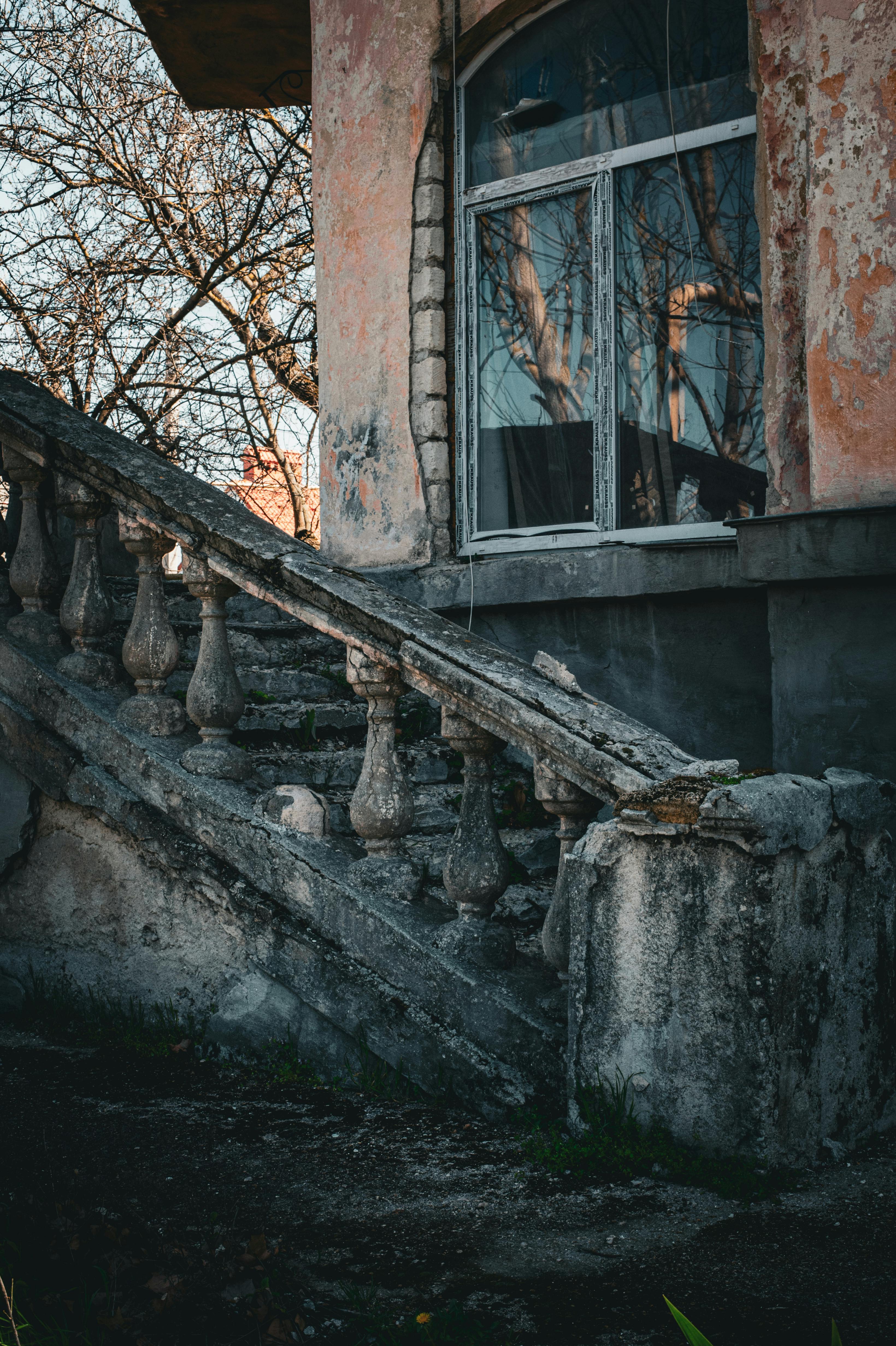 Gratis Una antigua escalera de piedra que conduce a un edificio desgastado con una ventana reflectante y árboles desnudos. Foto de stock