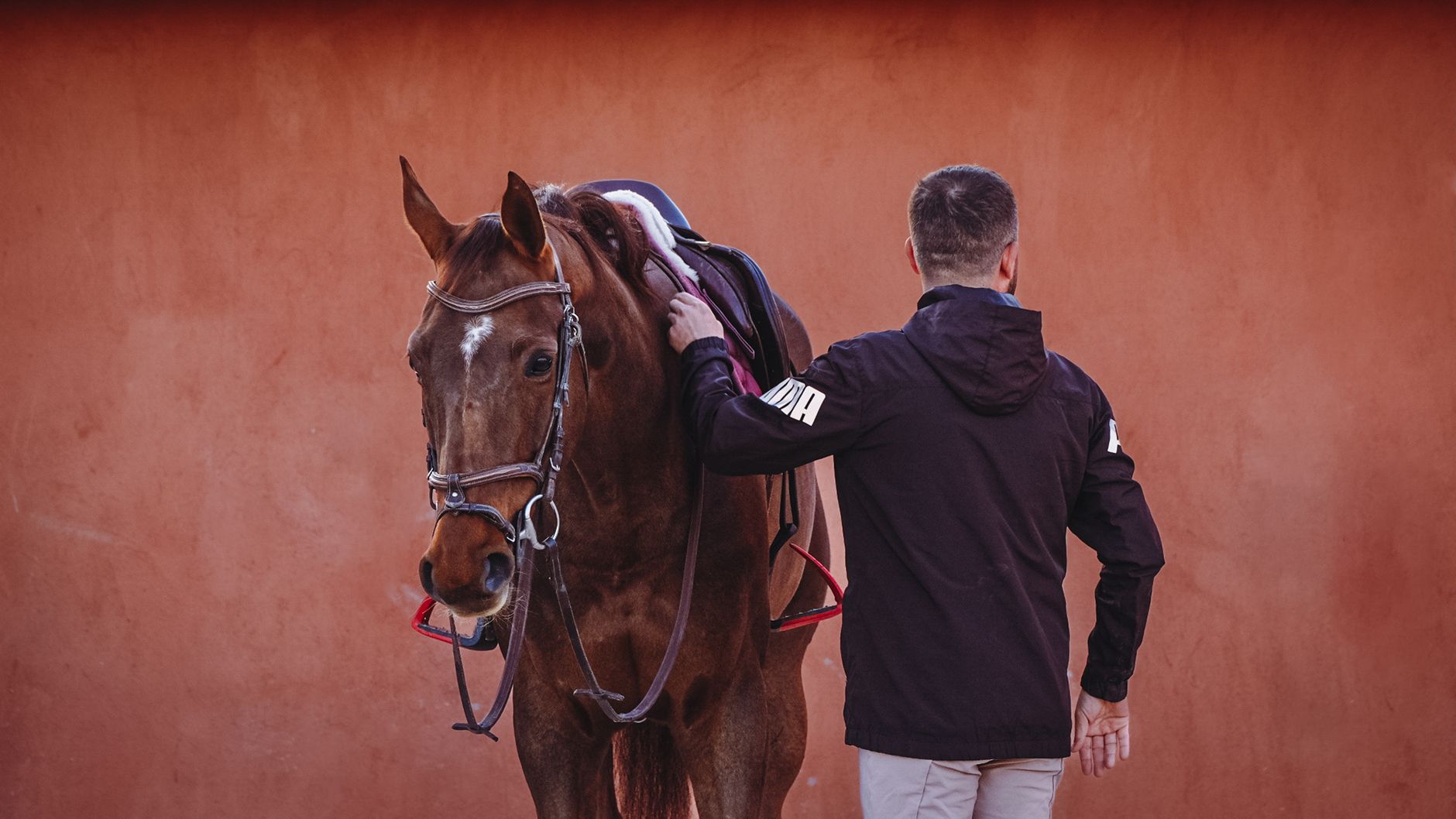 A man stands beside a brown horse in front of a terracotta wall, showcasing a tranquil moment.