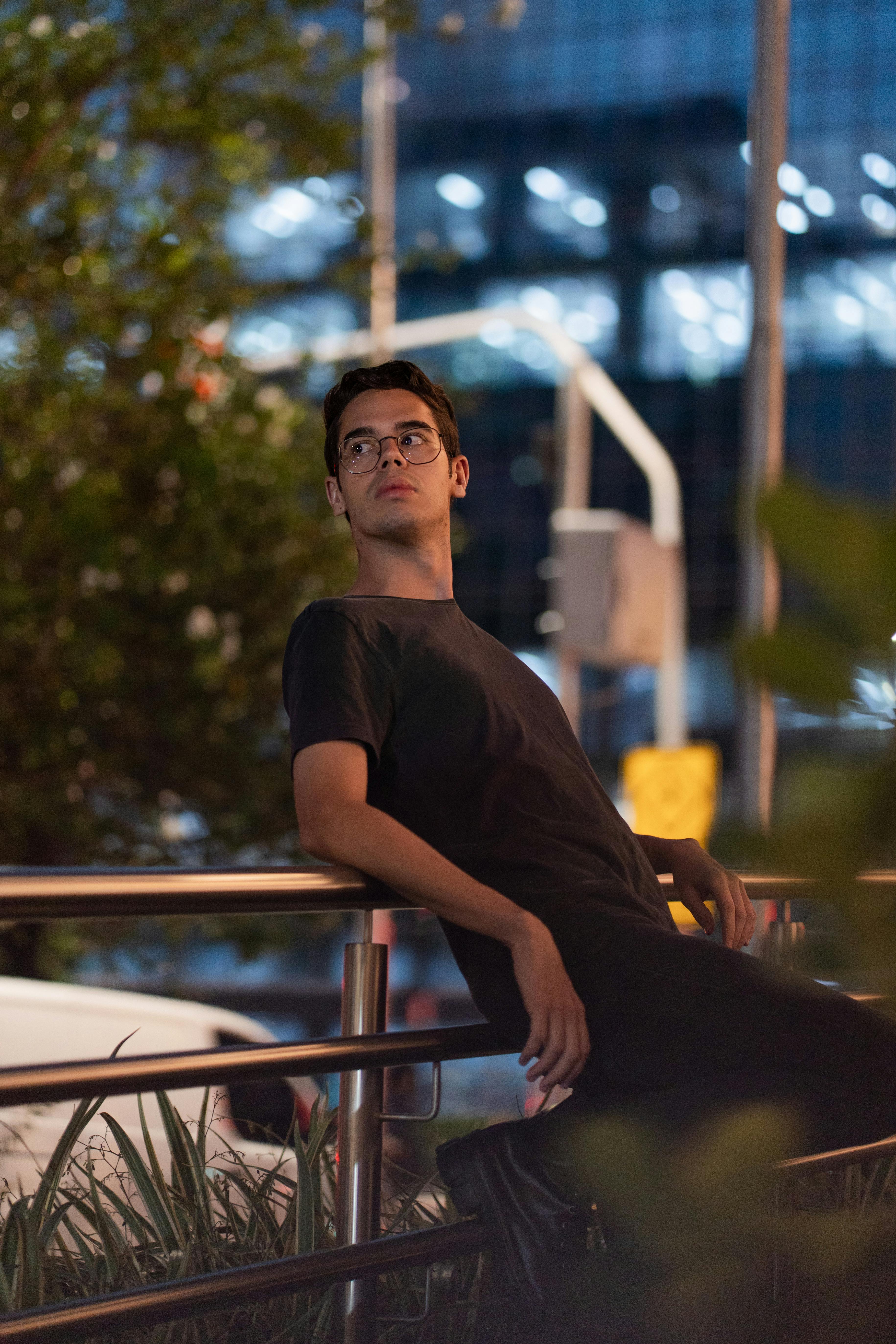 A young man wearing glasses leans pensively against a railing in an urban city setting at night.