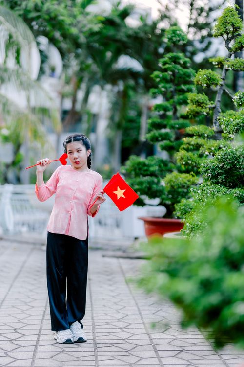 Free Young woman in traditional attire with Vietnamese flag, vibrant garden background. Stock Photo