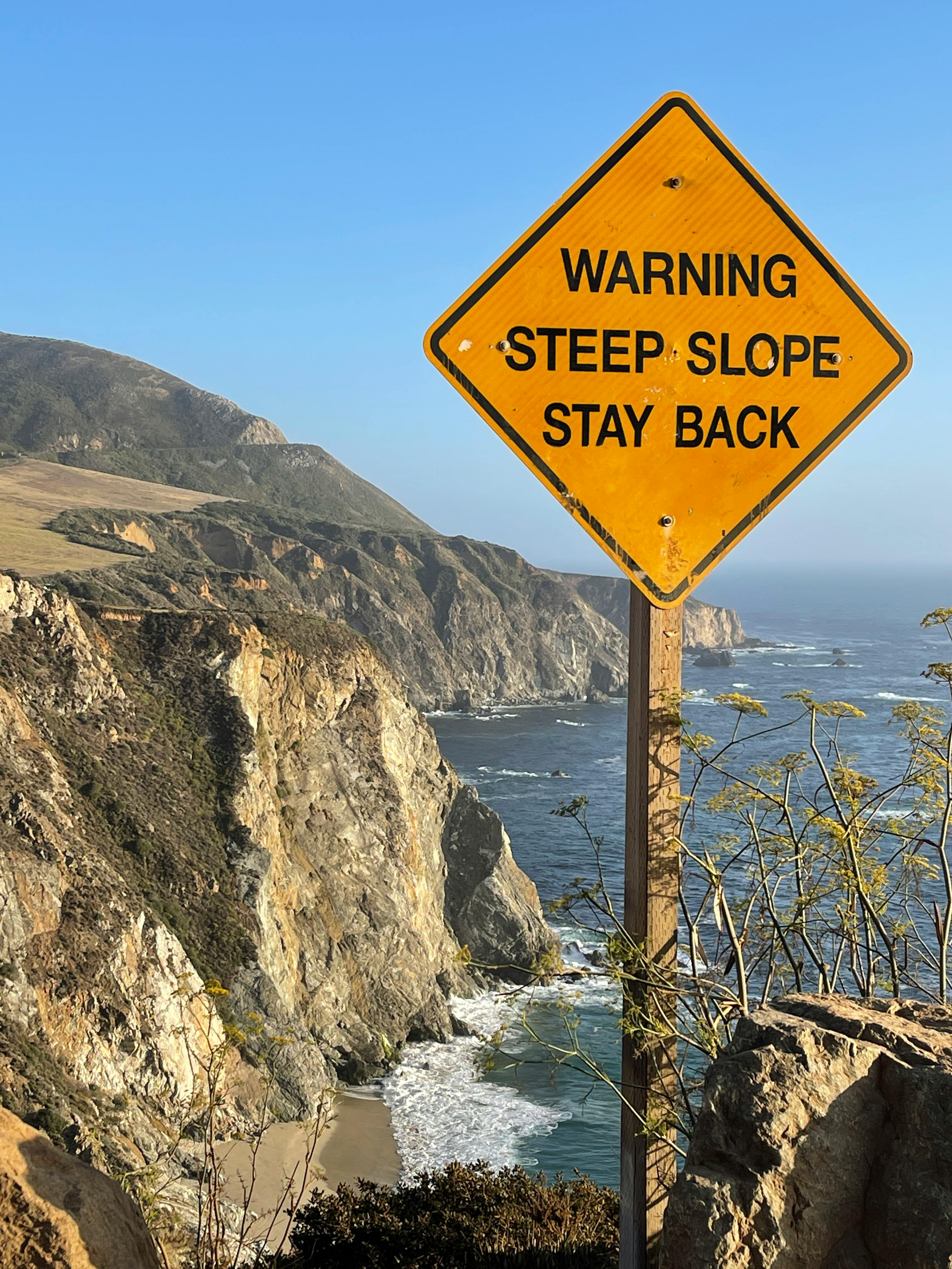 Yellow warning sign on steep cliffs of Big Sur coast, California.