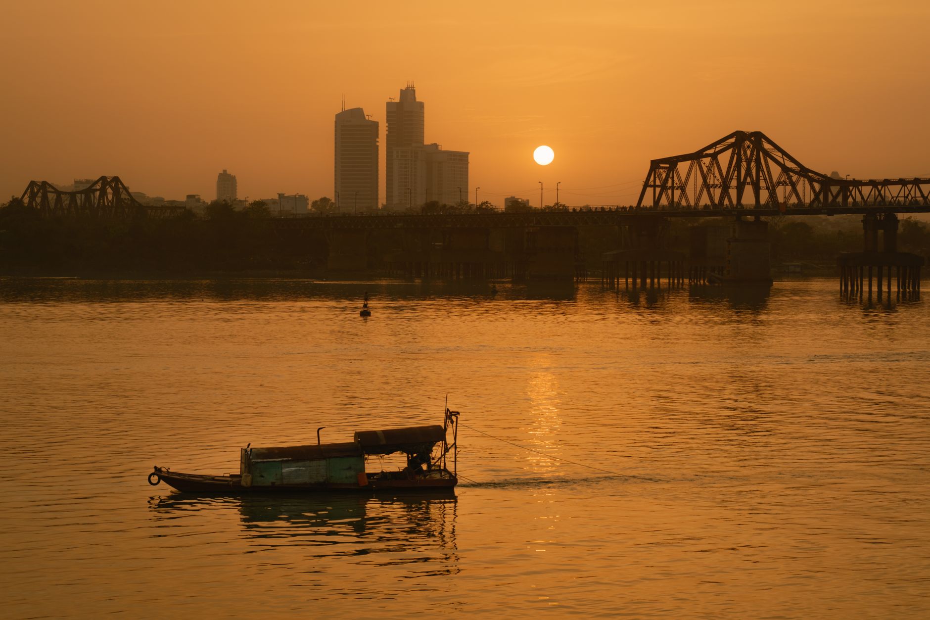 Tranquil sunset view of Long Bien Bridge in Hanoi with a boat on the river.