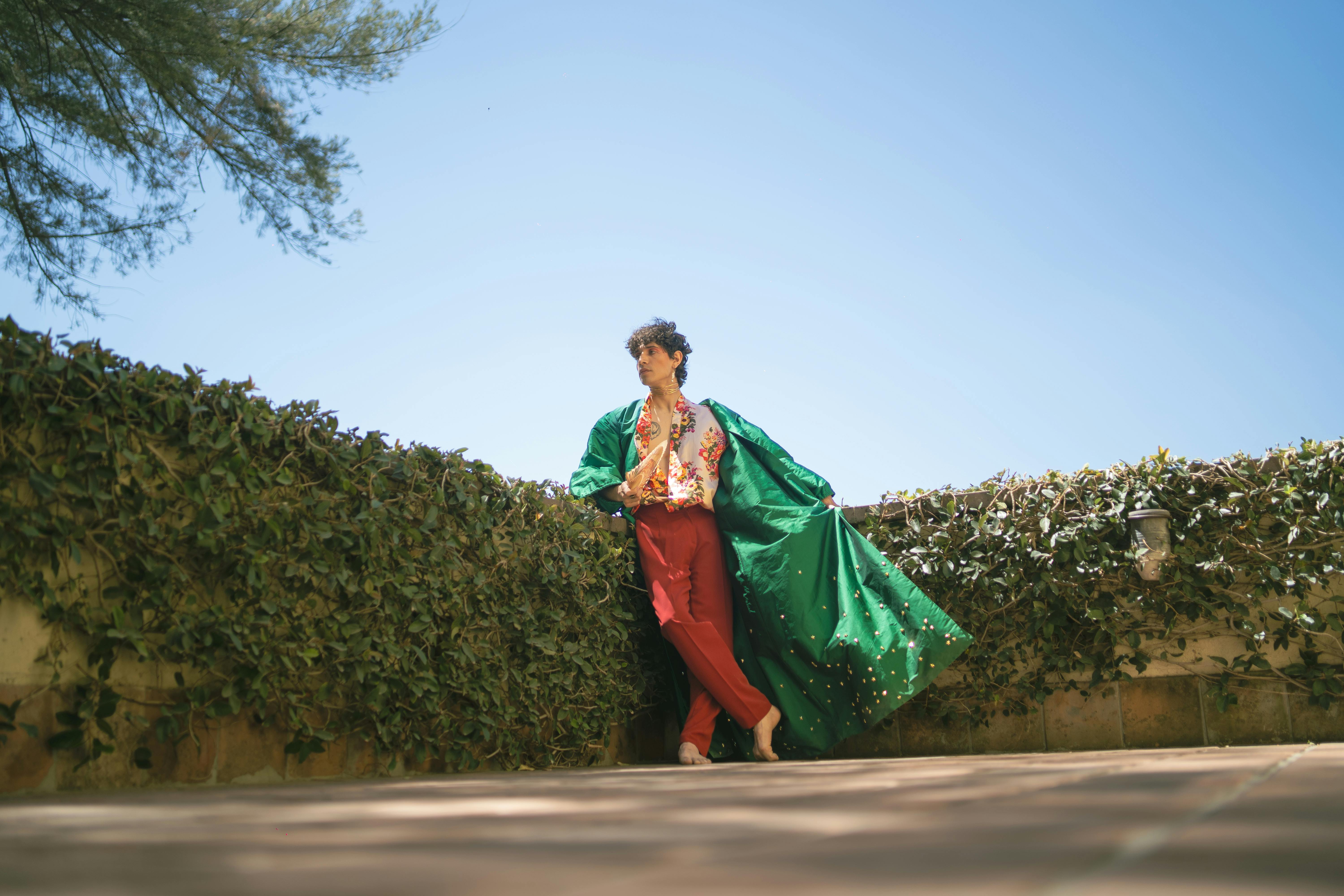 Stylish young man in vibrant attire posing outdoors against a lush green wall under a clear blue sky.