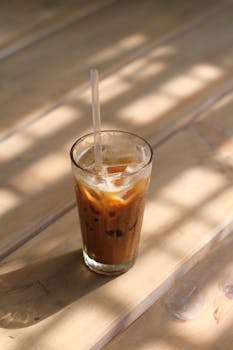 Glass of iced coffee on a wooden table, bathed in soft sunlight, creating a refreshing summer vibe.