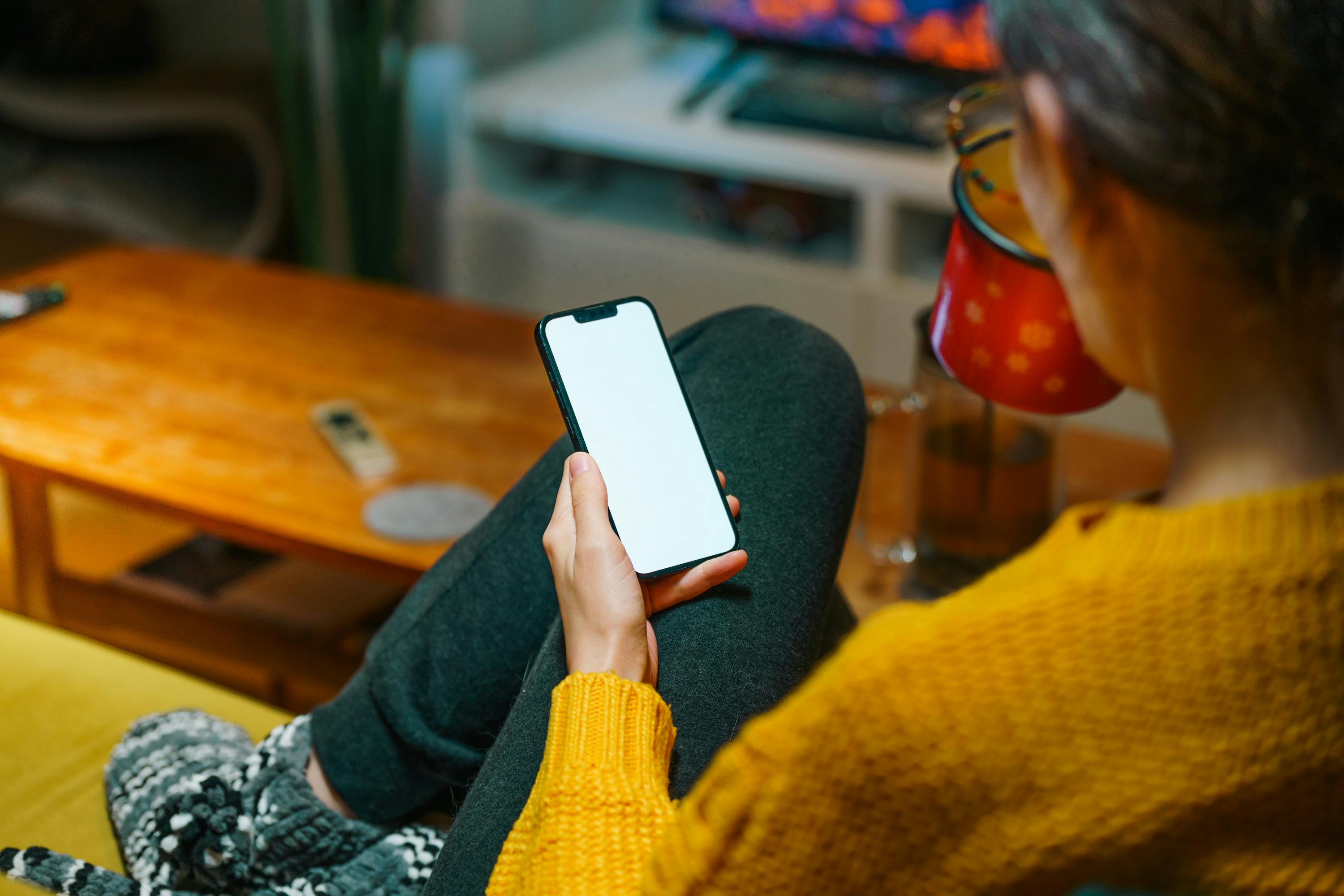 Gratis Una persona si rilassa sul divano con uno smartphone in mano, godendosi una calda serata invernale in casa. Foto a disposizione