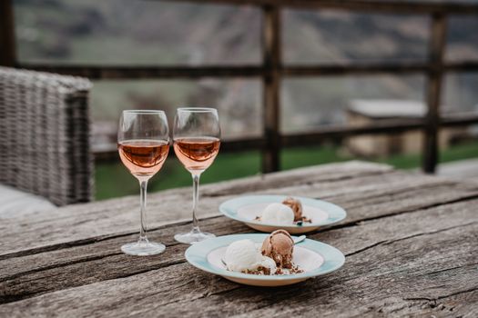 Rustic wooden outdoor table with two glasses of rosé wine and dessert plates featuring chocolate and vanilla ice cream.