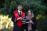 Two graduates in caps and gowns holding a clapperboard outdoors.