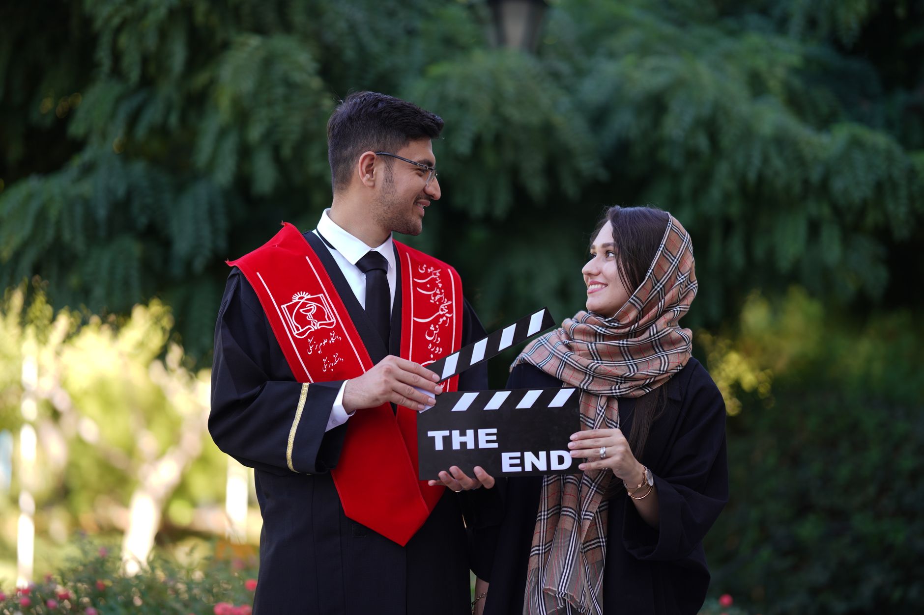 Two graduates in academic outfits holding a clapperboard reading 'The End', celebrating outdoors.