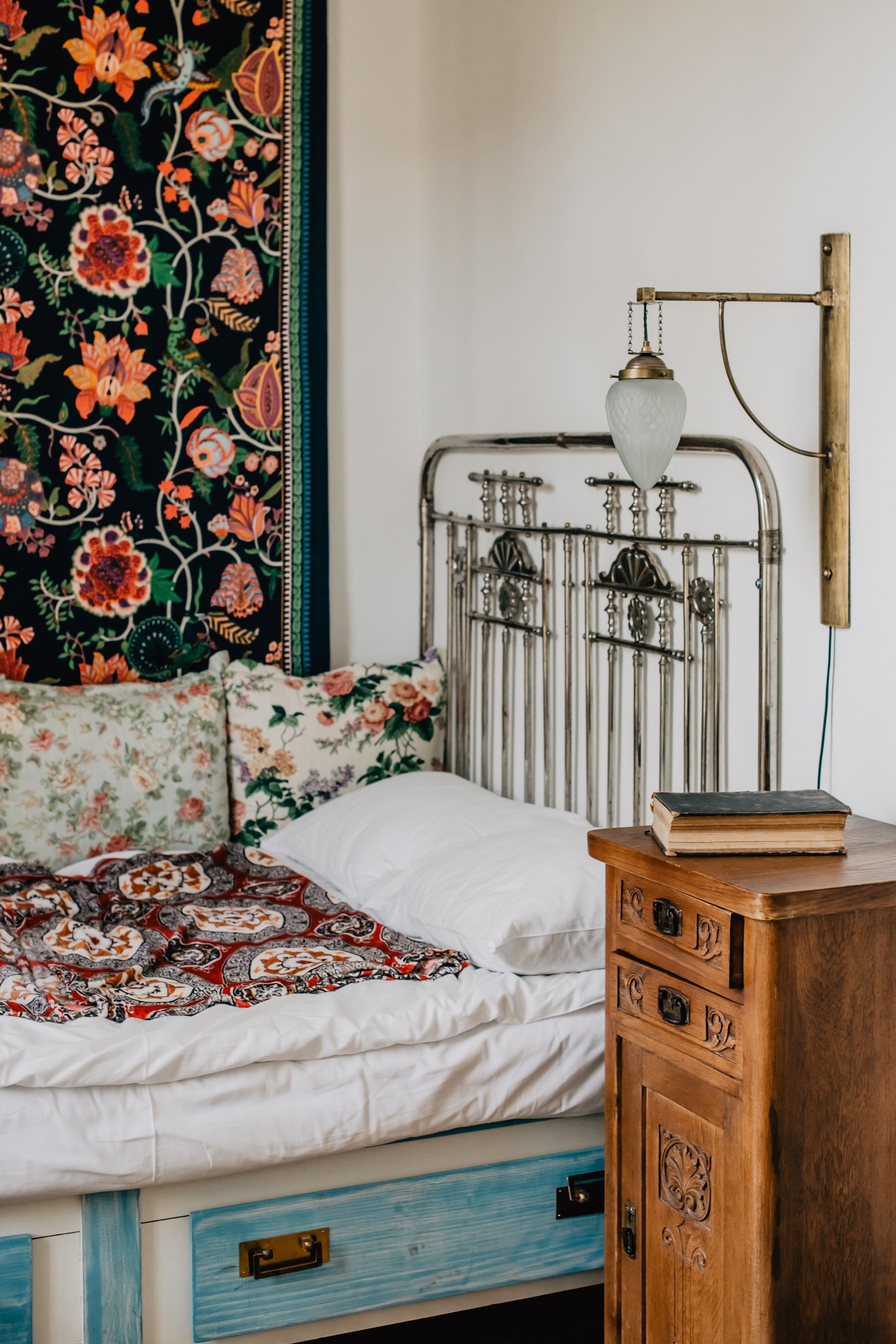 Vintage iron bed with floral tapestry, mixed patterned bedding and a wooden bedside chest