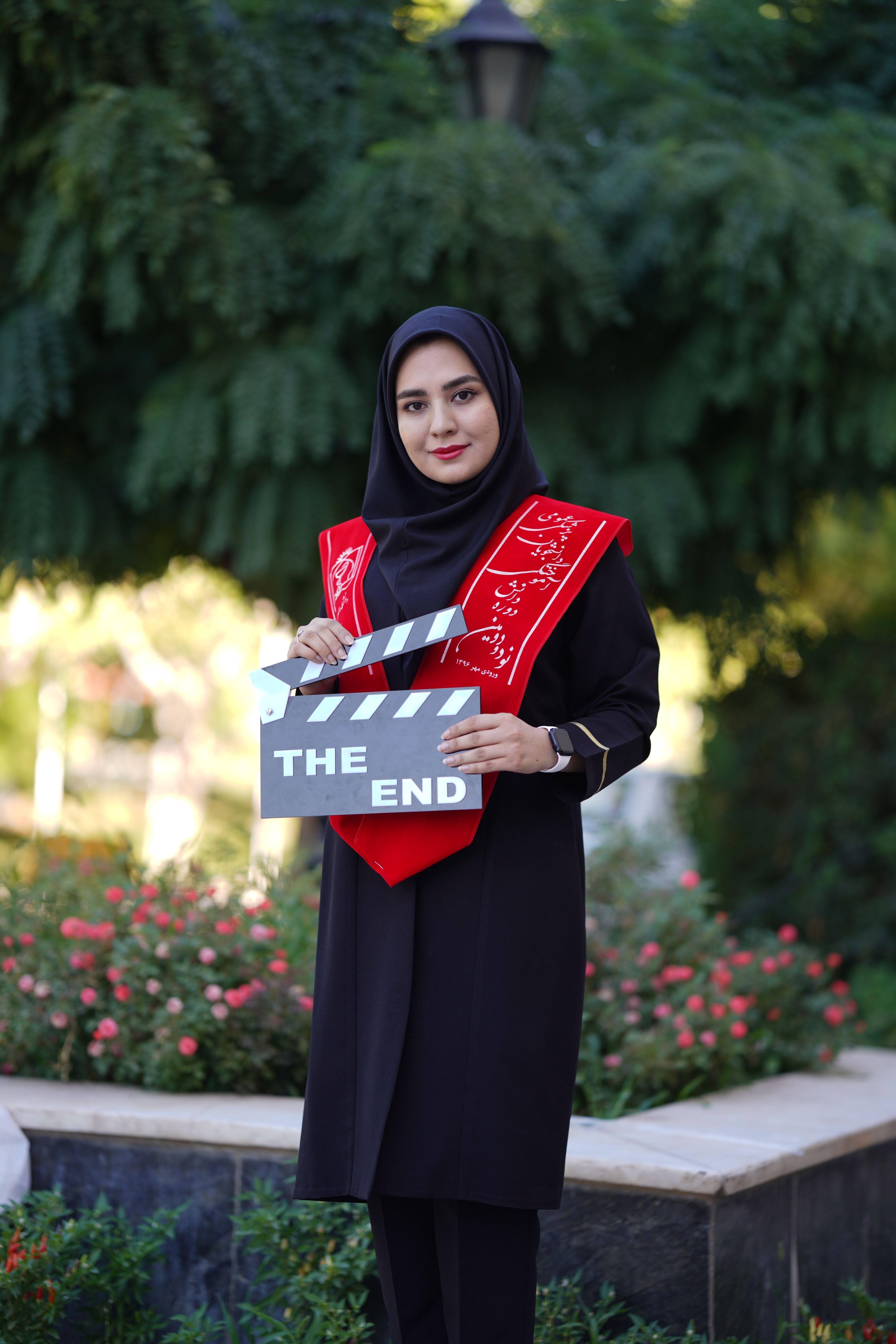 Young woman in a graduation stole holding a clapboard with 'The End' text outdoors.