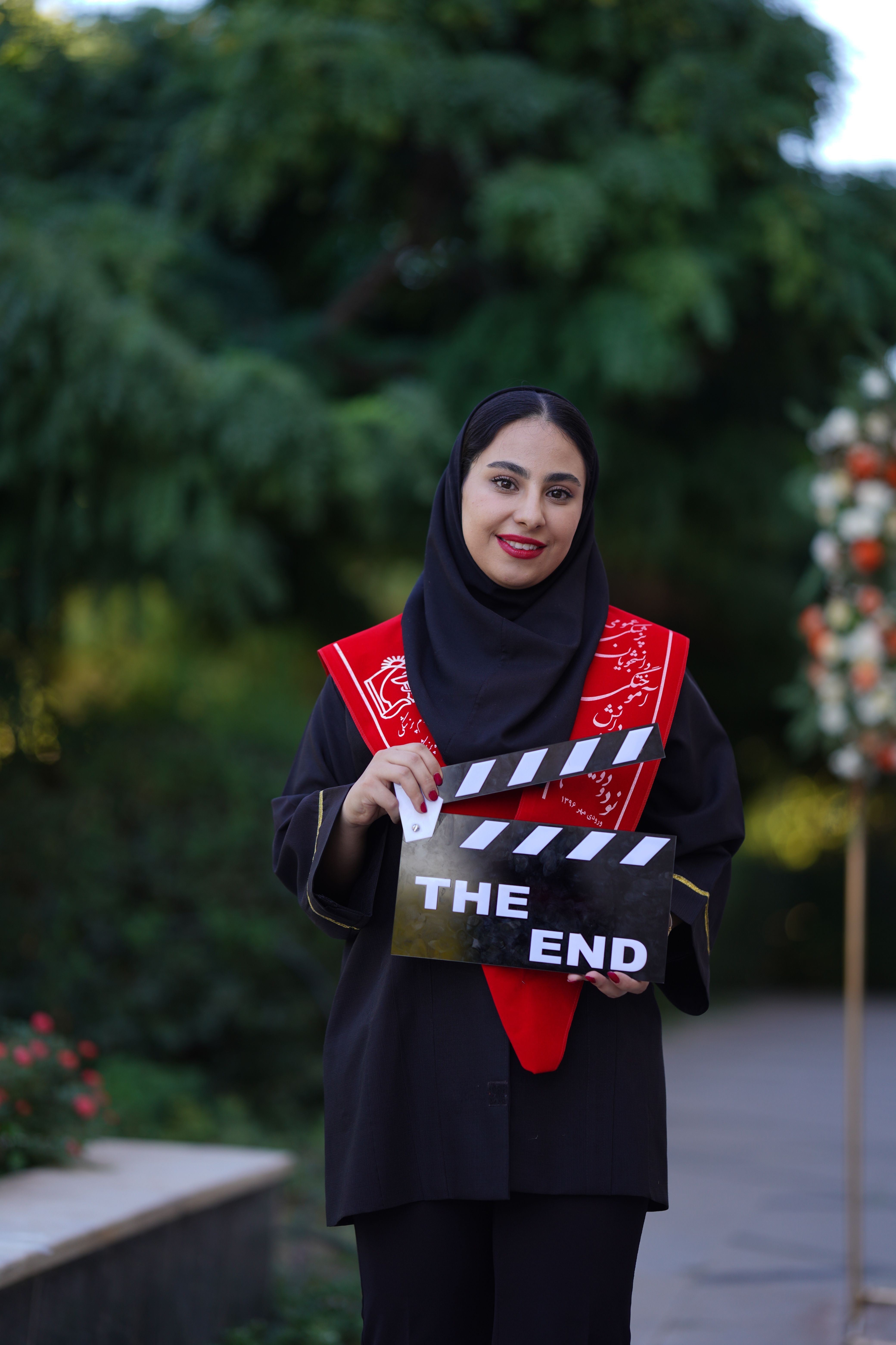 Young woman celebrating graduation outdoors with a ceremonial clapperboard.