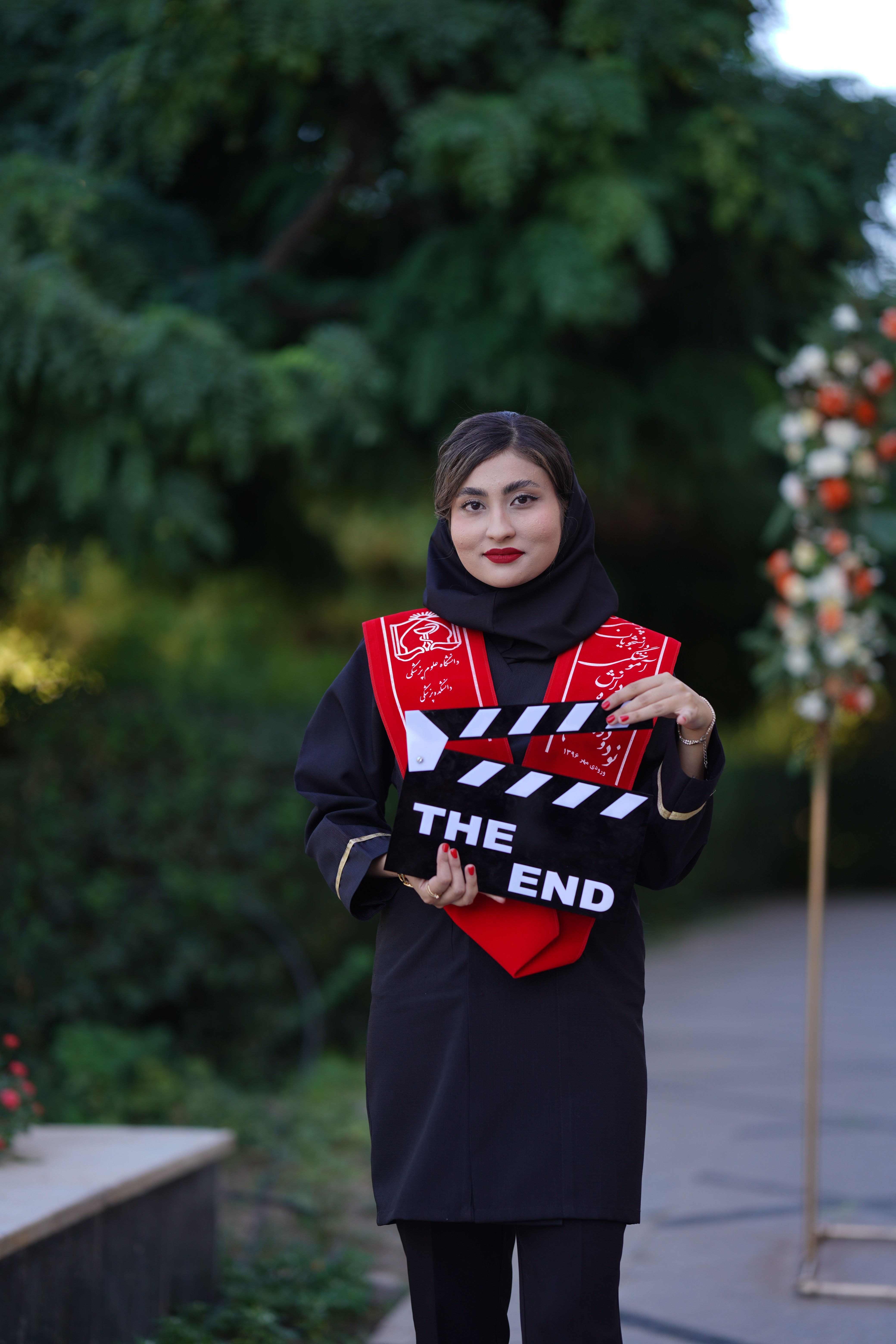 A woman in traditional Iranian attire holds a clapperboard marked 'THE END' at an outdoor graduation ceremony.