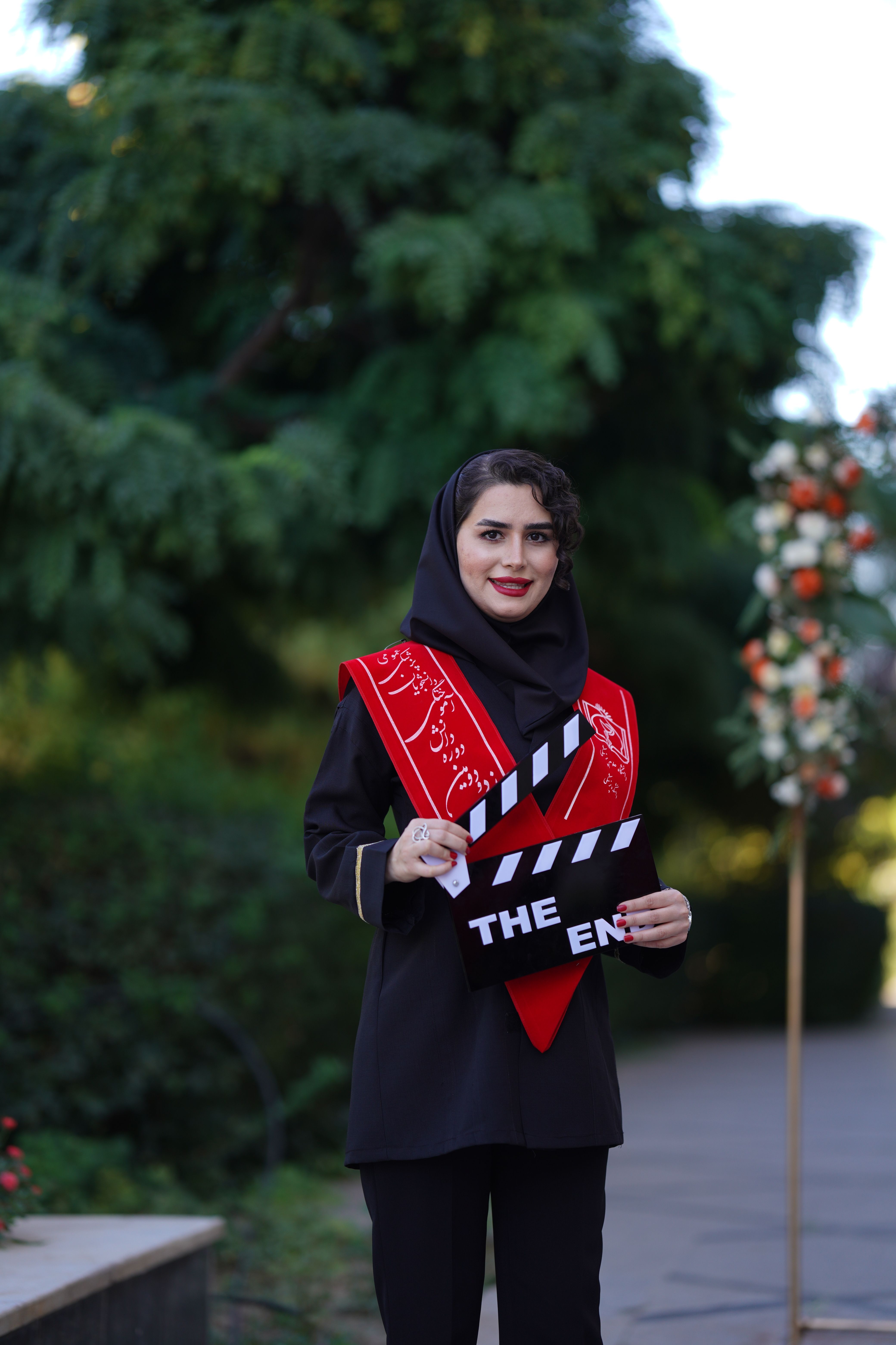 Smiling woman in hijab with clapperboard reading 'The End' in a park setting.