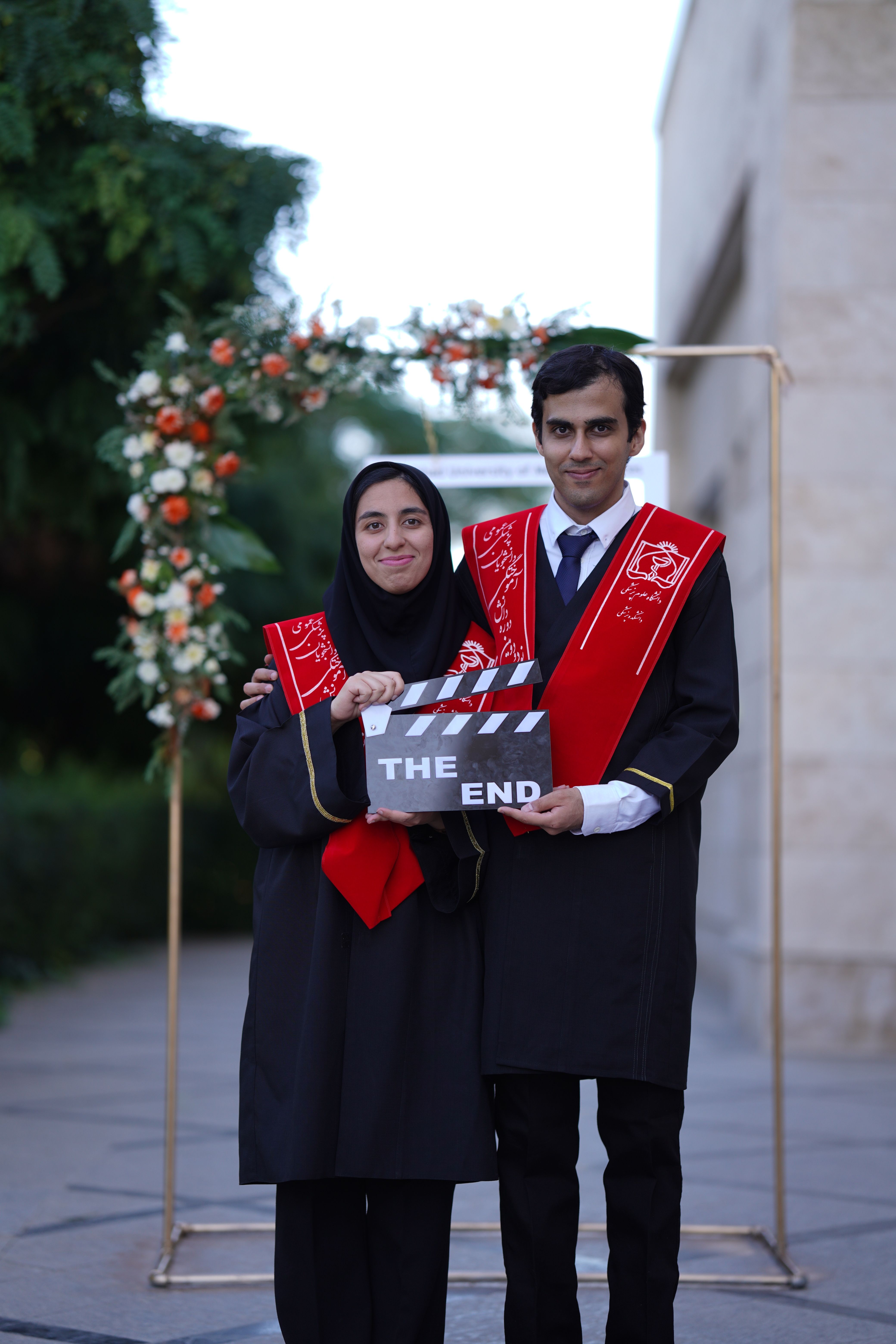Two graduates celebrate outdoors holding a 'The End' sign under a floral archway.
