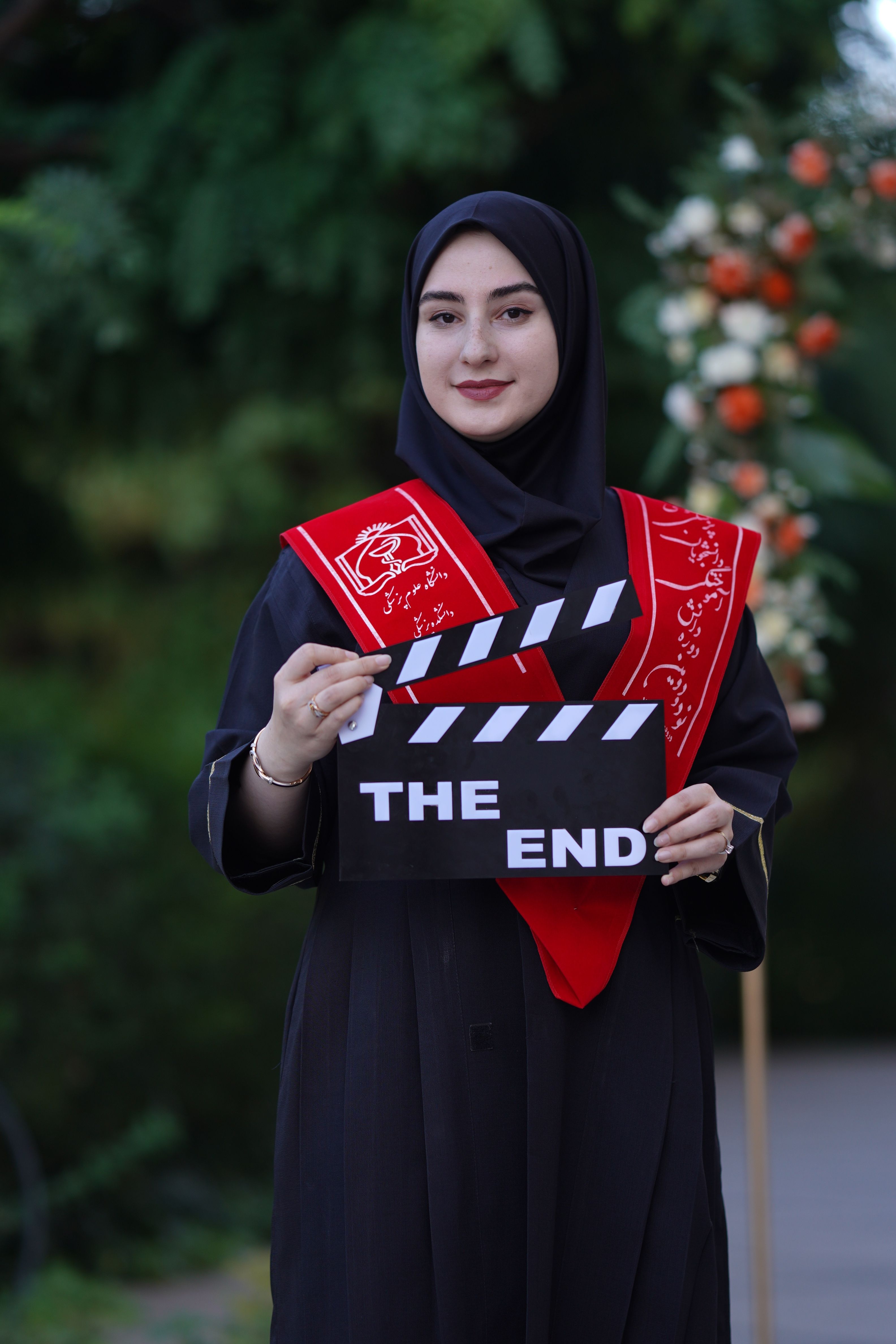 Young woman in hijab celebrating graduation with a clapperboard that reads 'The End' outdoors in a garden setting.