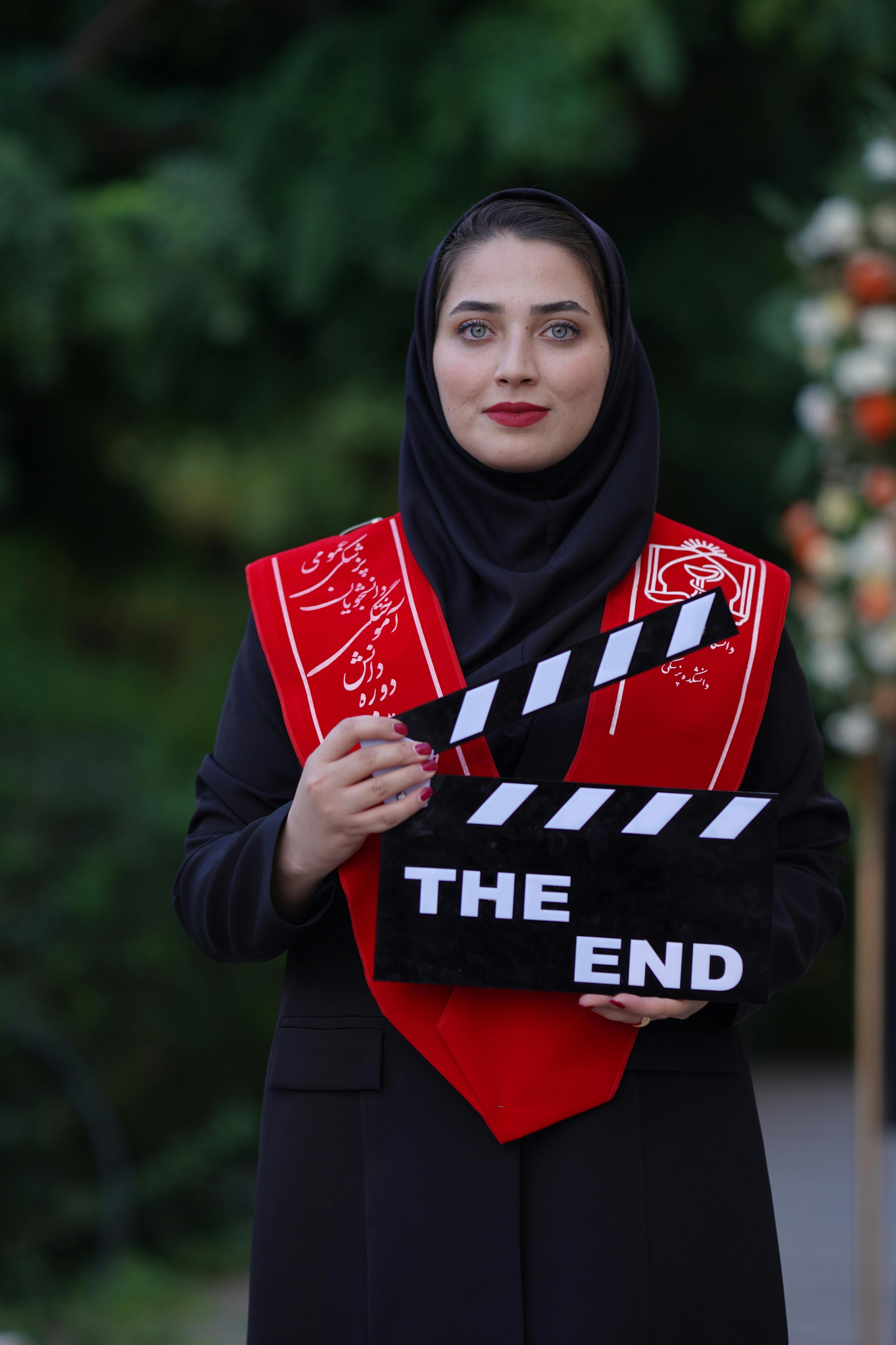 Young woman celebrating graduation in Iran, holding a clapperboard outdoors.
