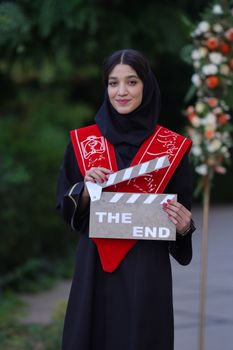 A young woman in graduation attire holding a clapboard saying 'The End'.