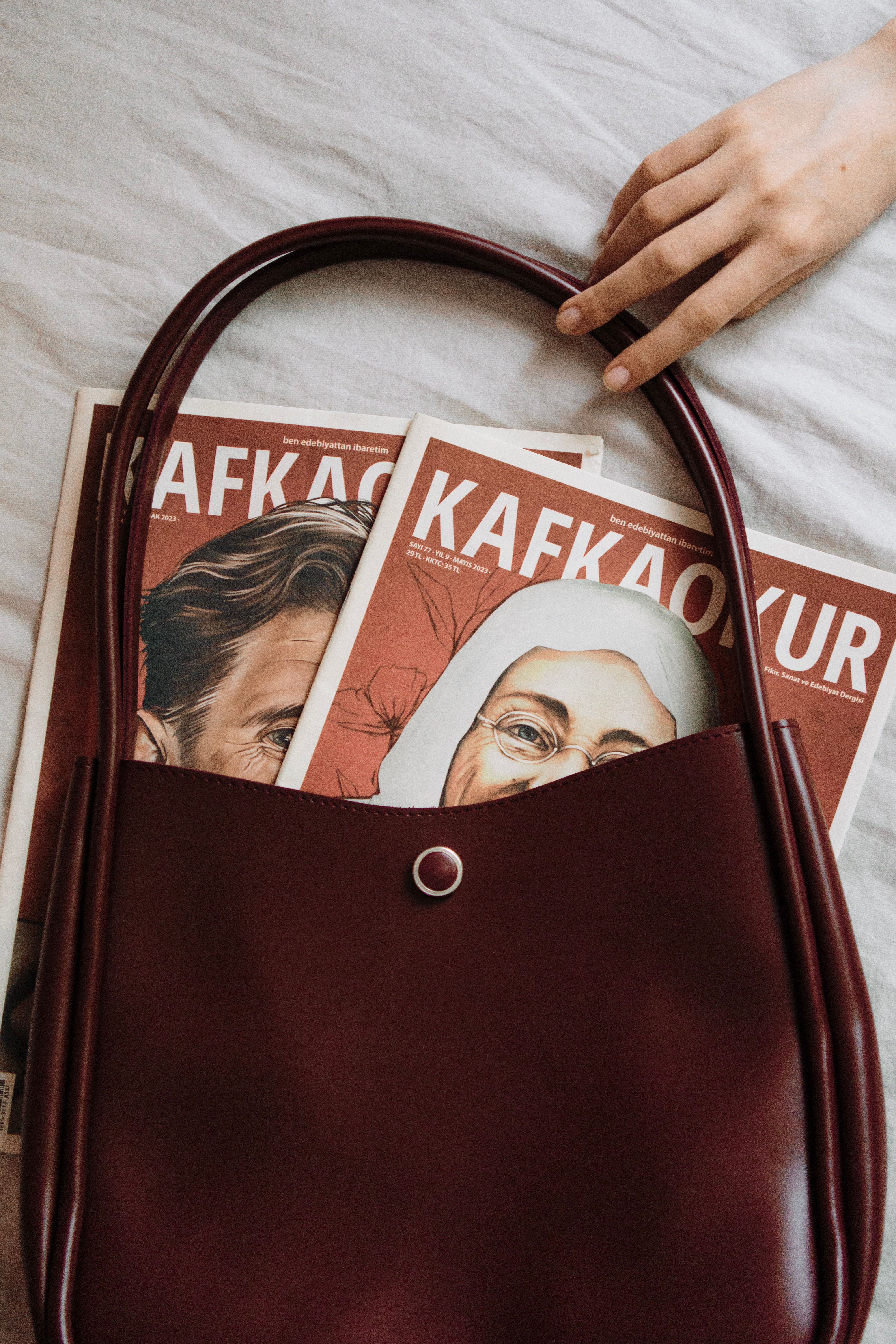 Closeup of a maroon bag with Kafka magazines partially in view, alongside a delicate hand.