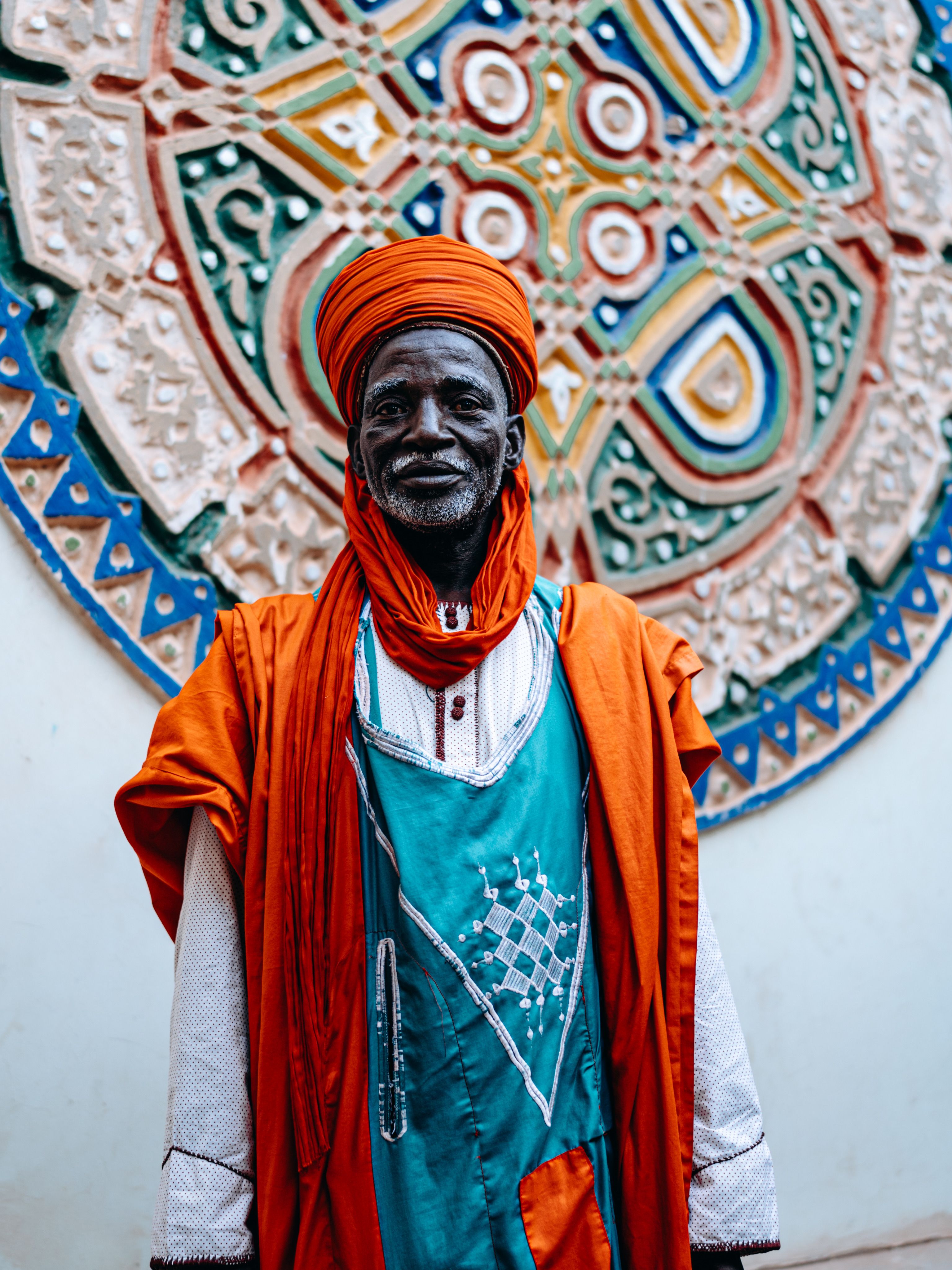 Portrait of an elder in traditional Nigerian attire, standing before decorative art.