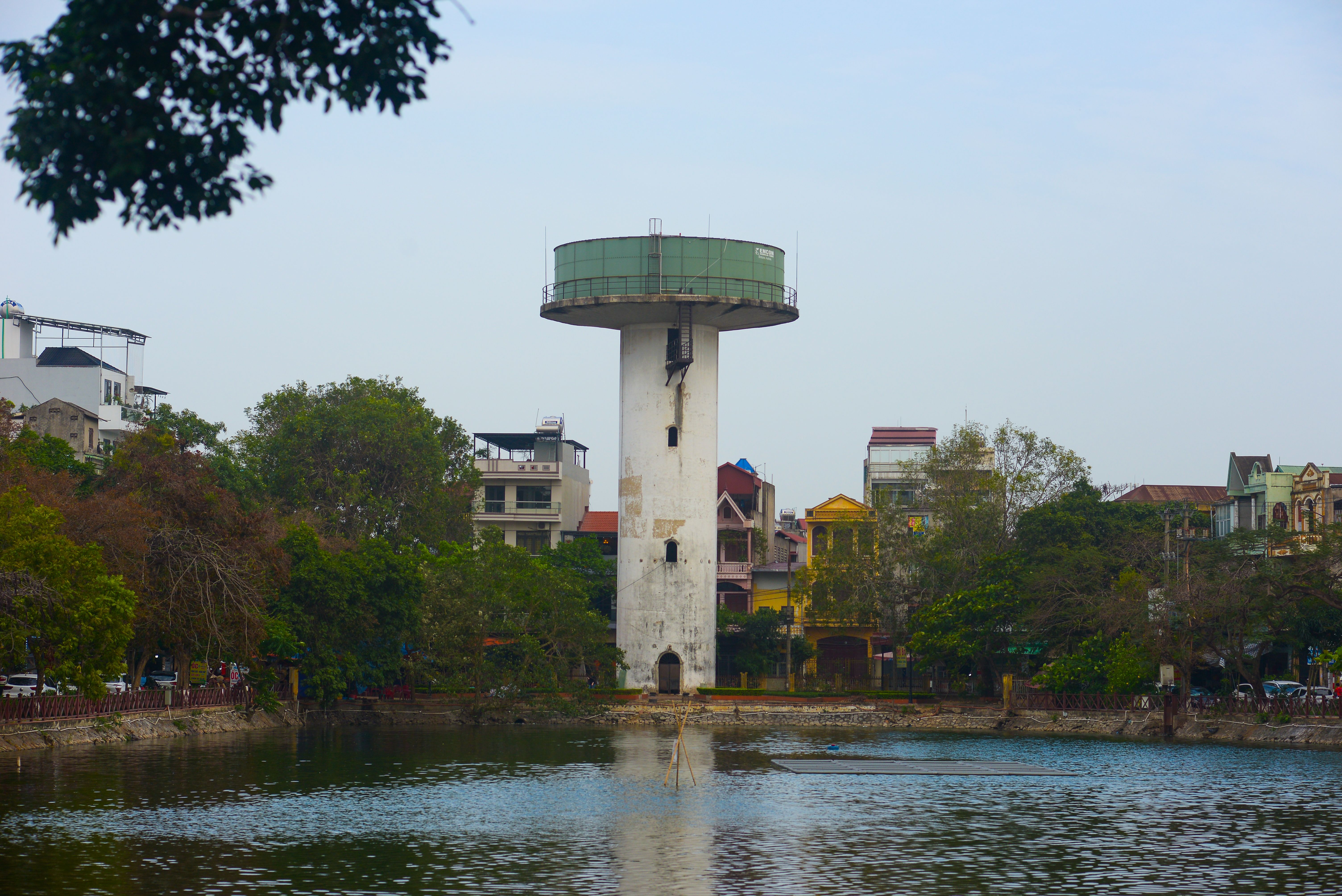 Torre de observação em meio a um parque urbano, refletida no rio ao seu lado, com casas e árvores ao fundo.