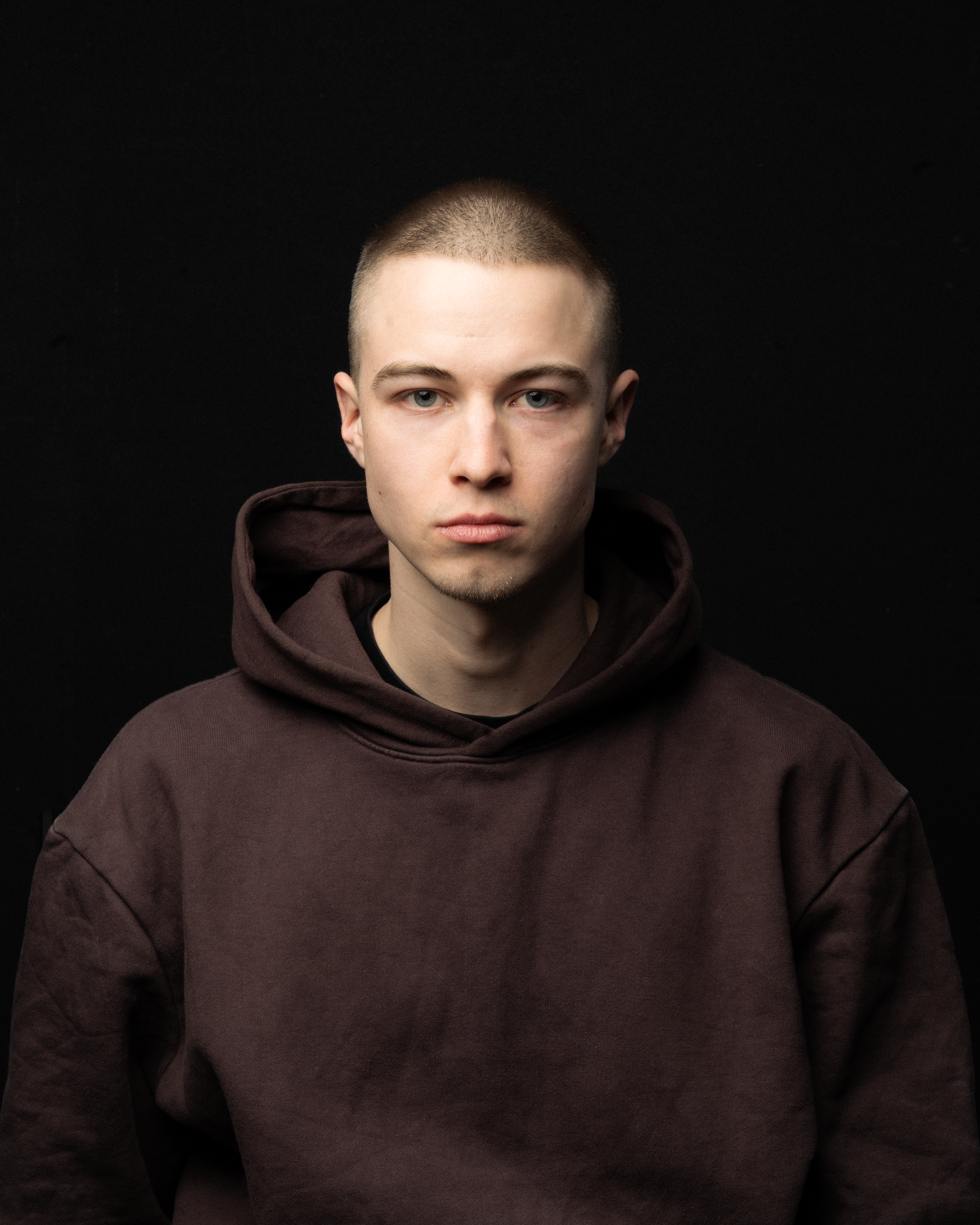 Close-up portrait of a young man wearing a brown hoodie against a black background.